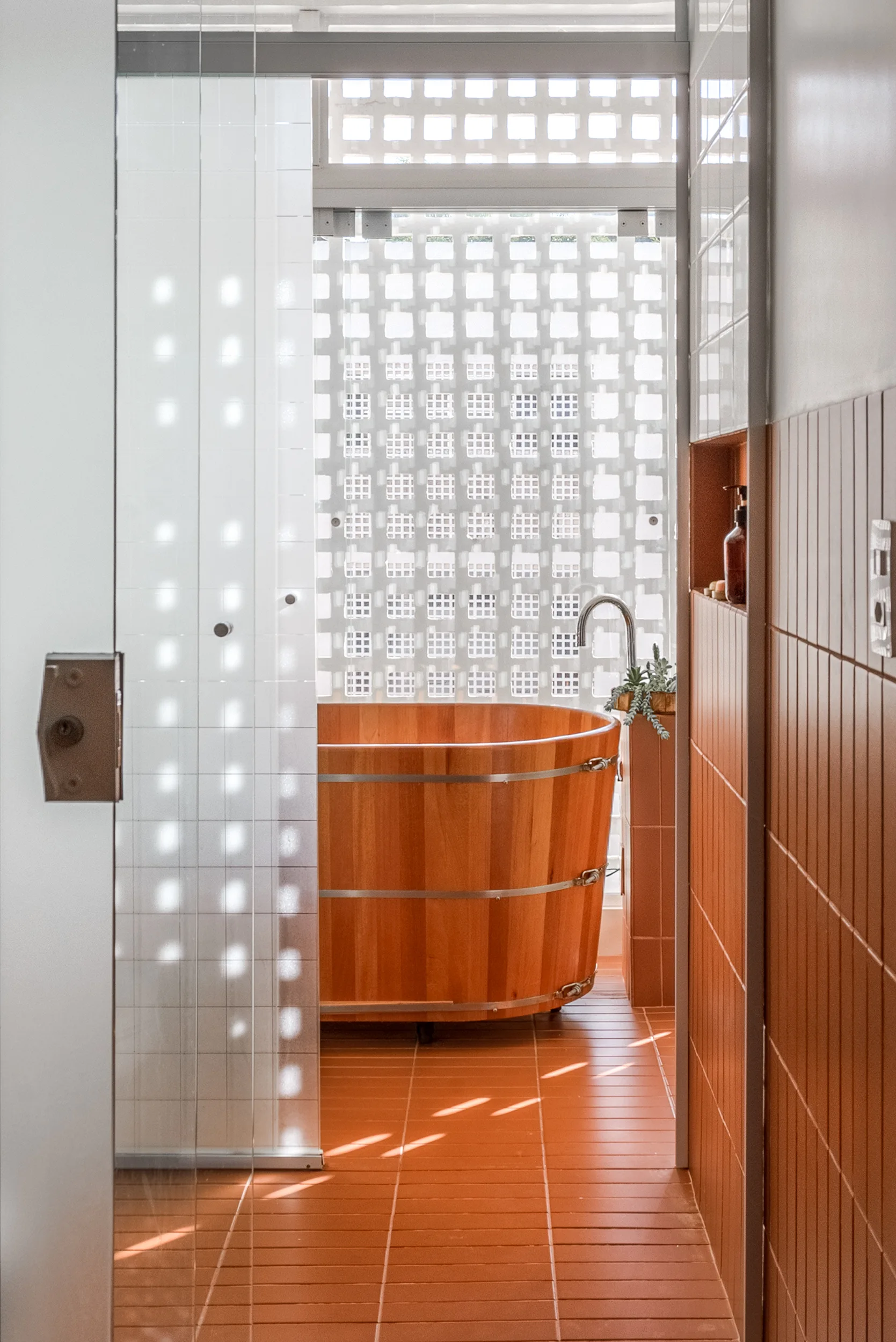 A bathroom featuring a round wooden soaking tub set against a white breeze block wall, surrounded by warm terracotta floor and wall tiles.