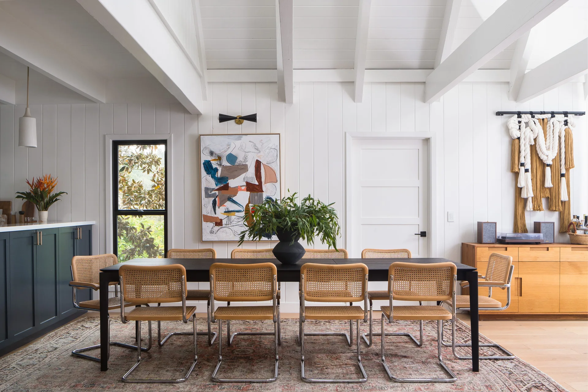 A bright dining room features a black wood table with cane-webbing chairs set on a patterned rug under white painted ceiling beams.