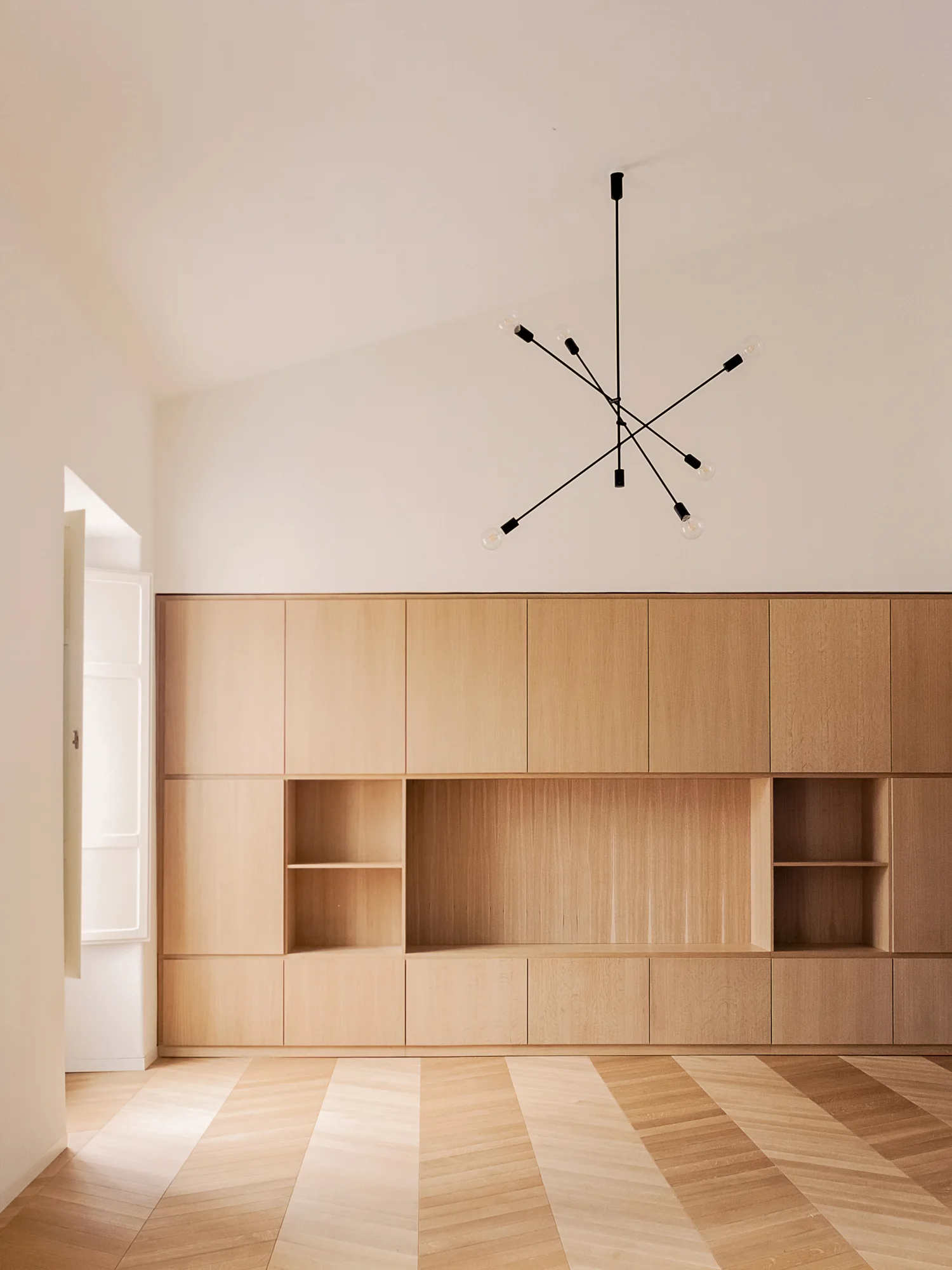 A minimalist room featuring floor-to-ceiling light wood cabinetry, herringbone patterned wood flooring, and a black multi-arm ceiling pendant.