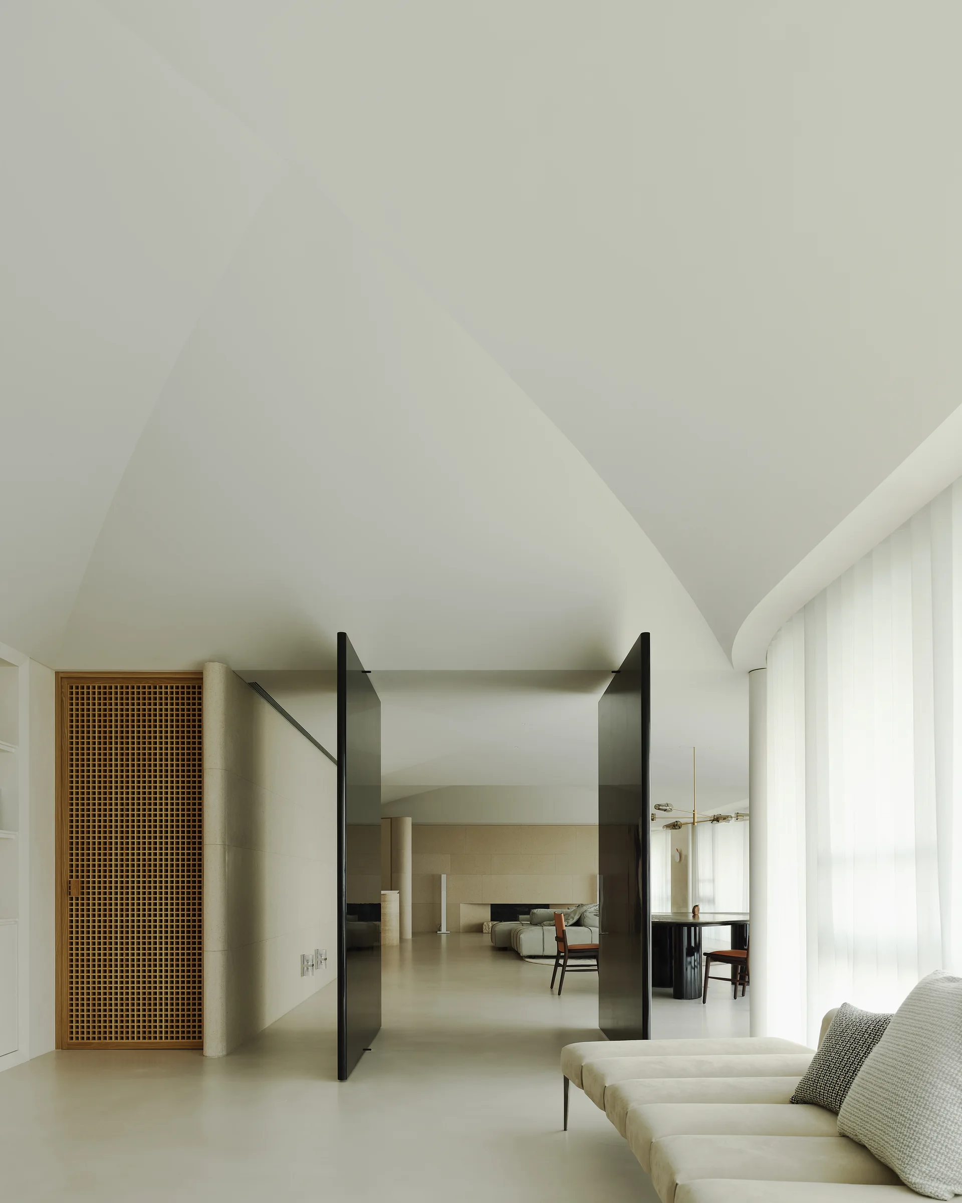 A minimalist cream living room features tall black pivot doors opening to a dining area under a vaulted ceiling, with sheer curtains on the right.