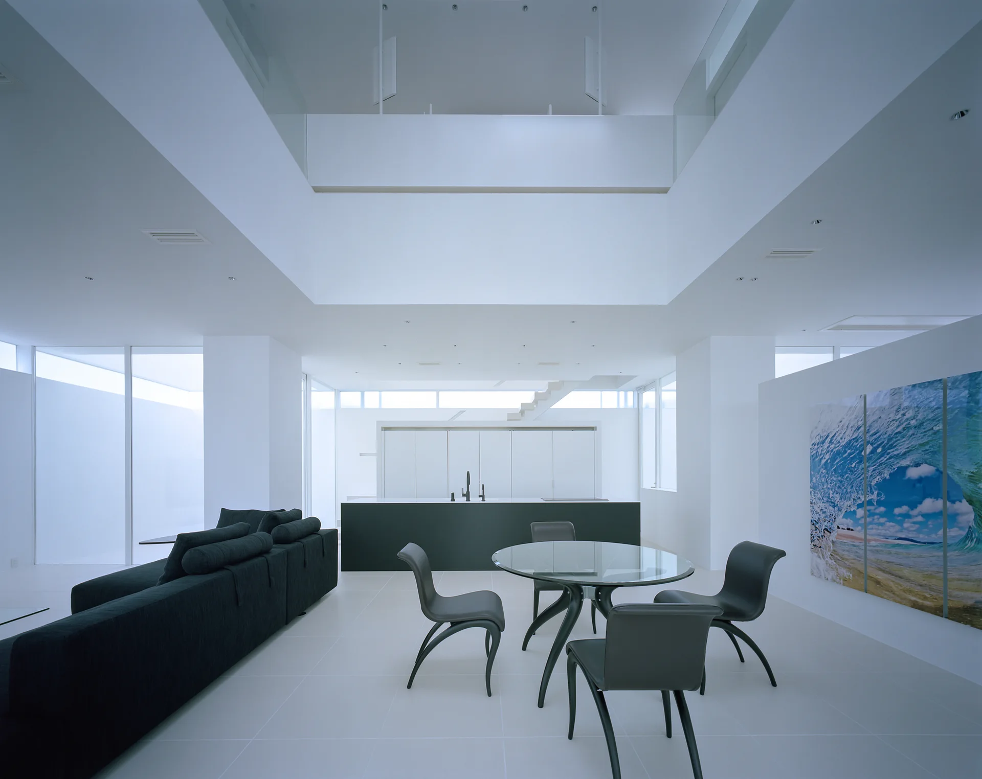 A minimalist open-plan living space features a dark sofa, a glass-topped dining table, and a black kitchen island under a soaring white ceiling.