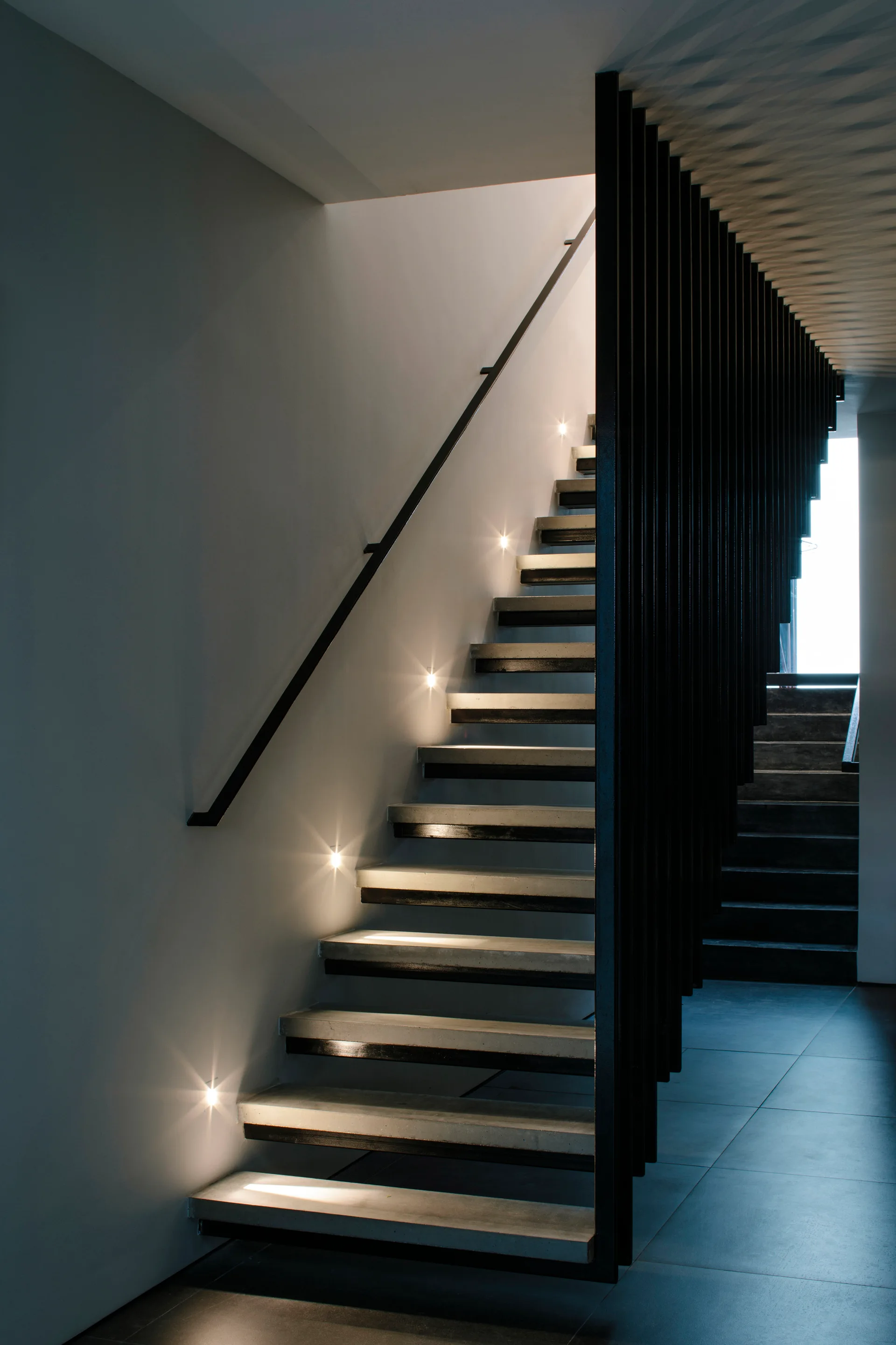 A modern minimalist staircase with floating concrete treads, illuminated by recessed wall spotlights and bordered by a vertical black metal slat screen.