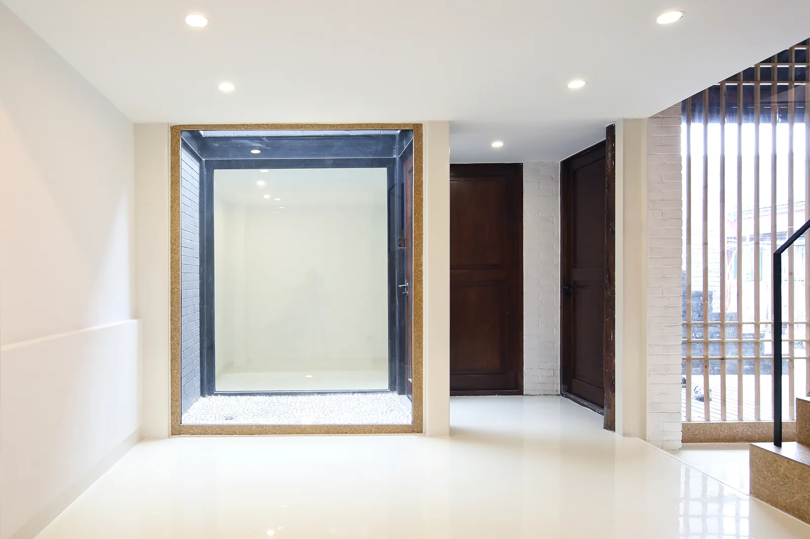 A minimalist entryway features a large glass light well filled with white gravel, glossy white floors, and two dark wood doors.