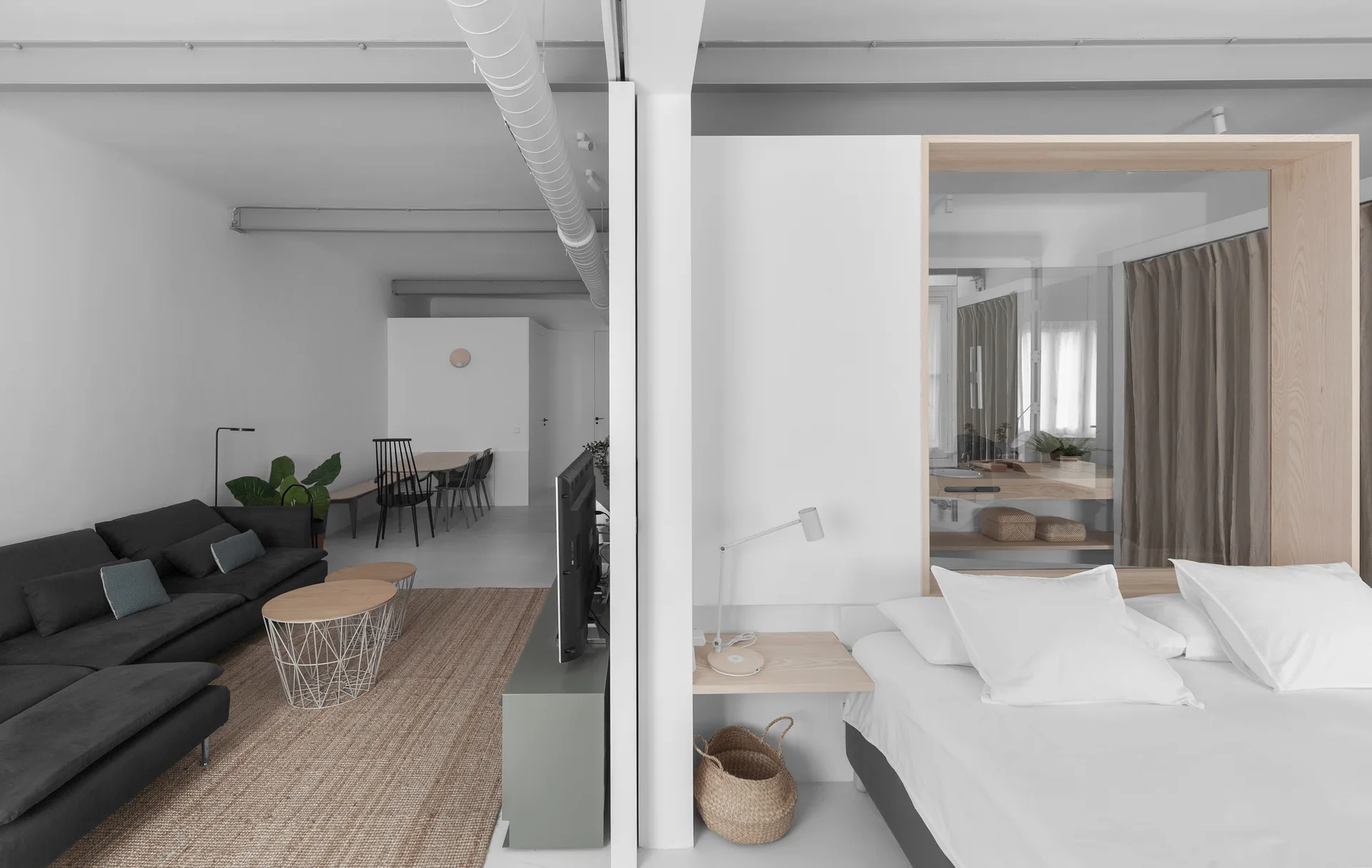 A central white partition separates a grey living room with a woven rug from a bedroom featuring white bedding and a wood-framed glass window.