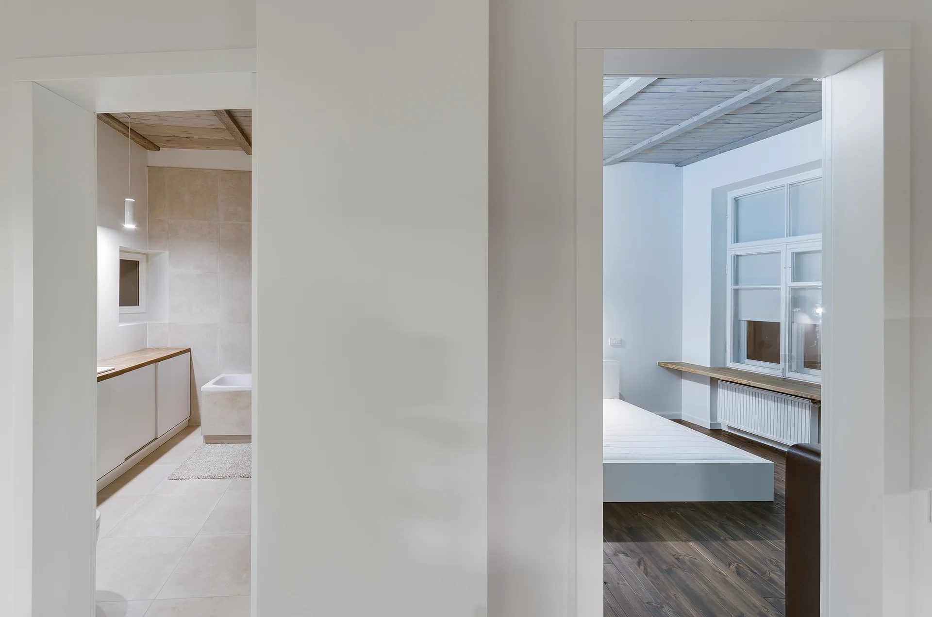A view through two white-framed doorways showing a tiled bathroom with a wood counter on the left and a minimalist bedroom with dark wood floors on the right.