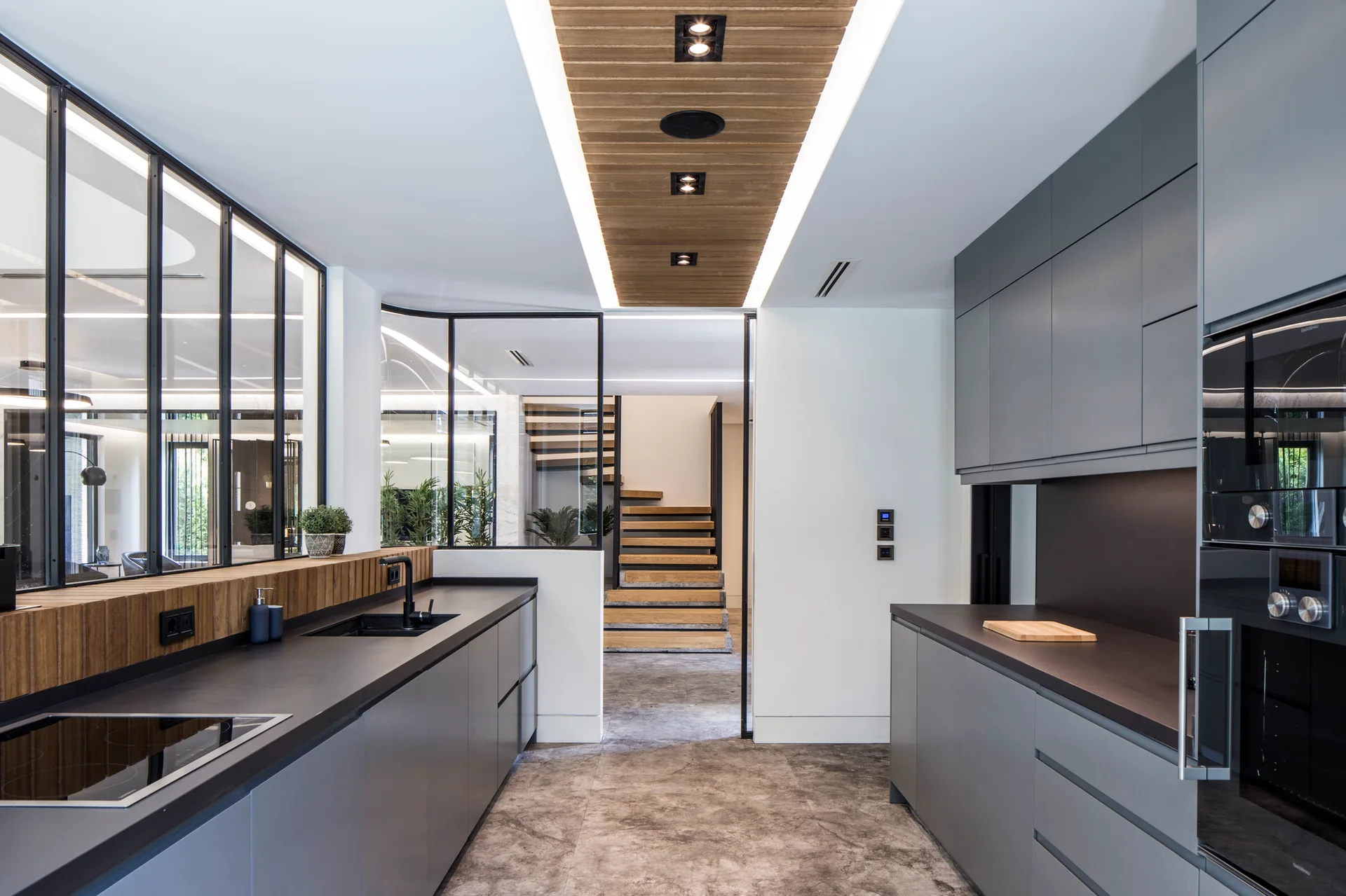 A modern galley kitchen features matte gray cabinets, dark countertops, a wooden slatted ceiling inlay, and a black-framed glass partition.
