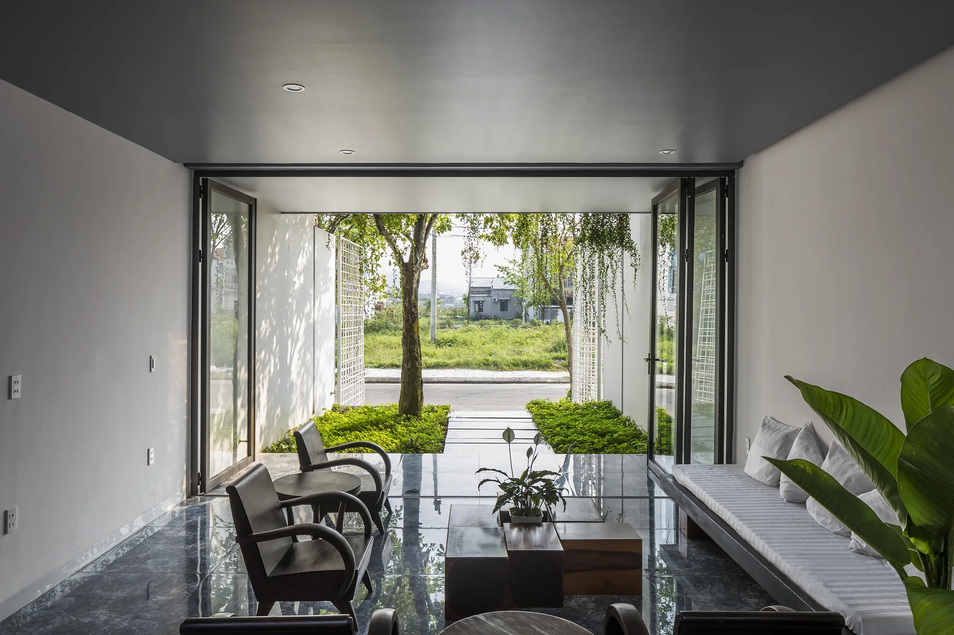 A modern living room featuring dark polished floor tiles, dark wood armchairs, and folding glass doors that open to a small green courtyard.