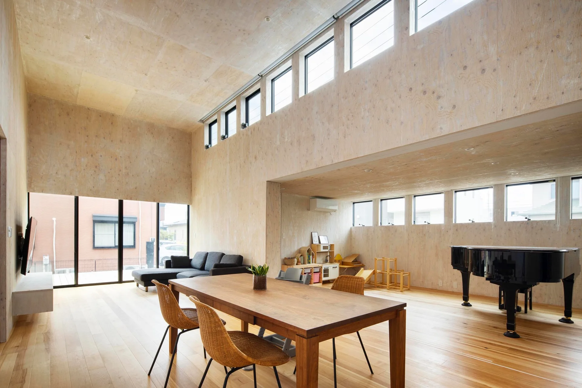 An open-plan residential interior with light plywood walls and ceiling features a wooden dining table, a grey sofa, and a black grand piano.