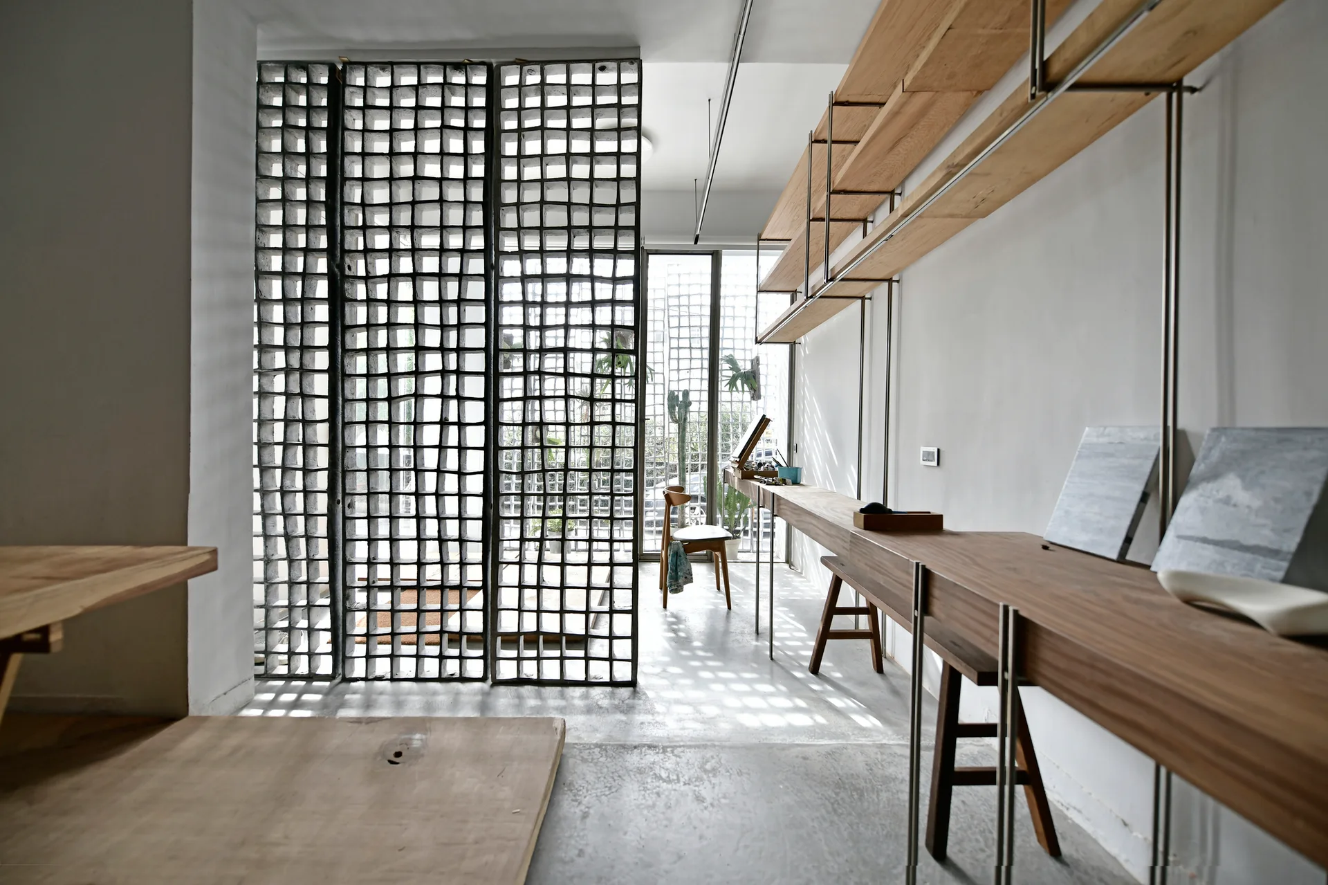 A minimalist workspace featuring a long wall-mounted wooden desk, metal shelving, and a large concrete grid screen partition next to a window.