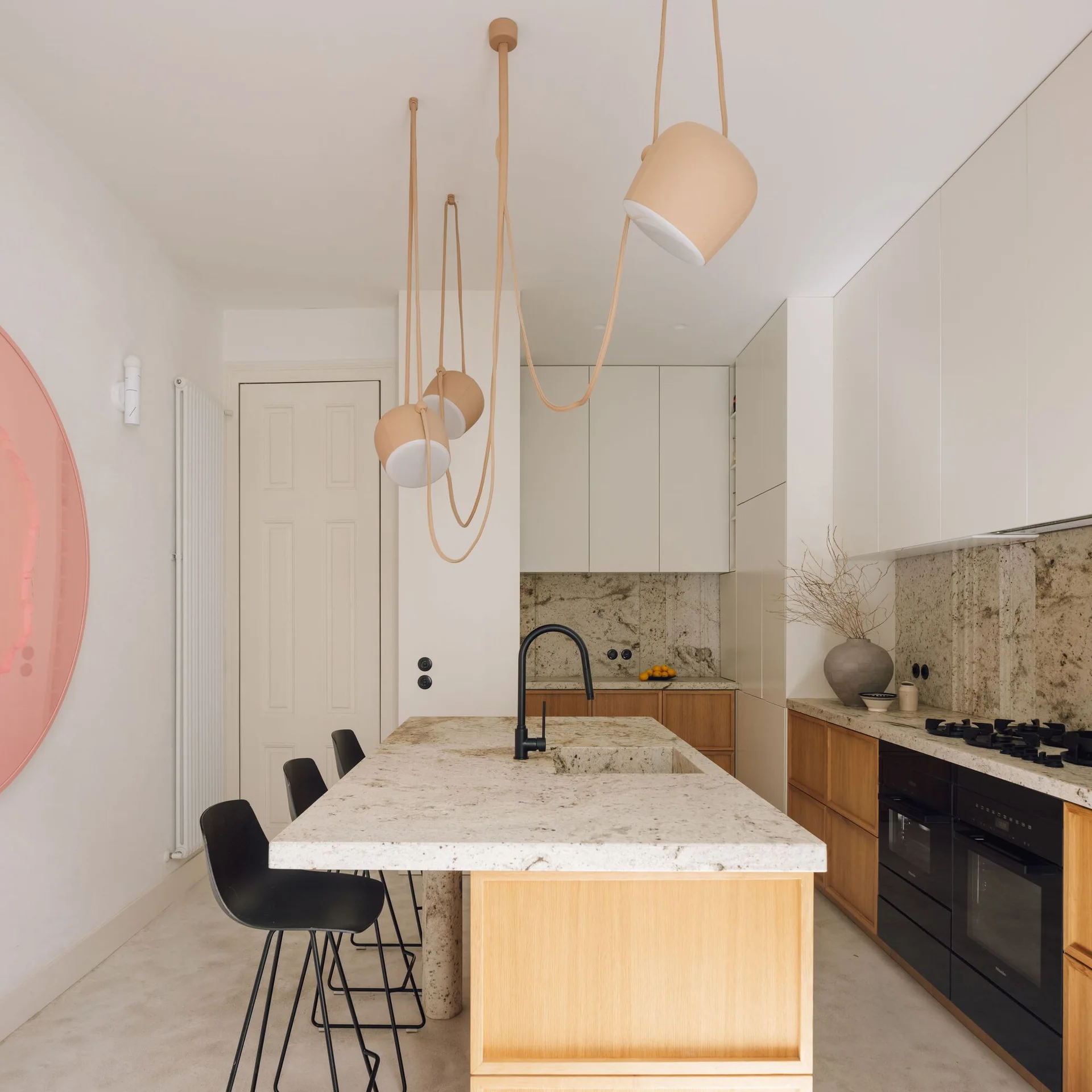 A minimalist kitchen features a stone-topped island with black stools, wood cabinetry, and a multi-light peach pendant fixture draped from the ceiling.