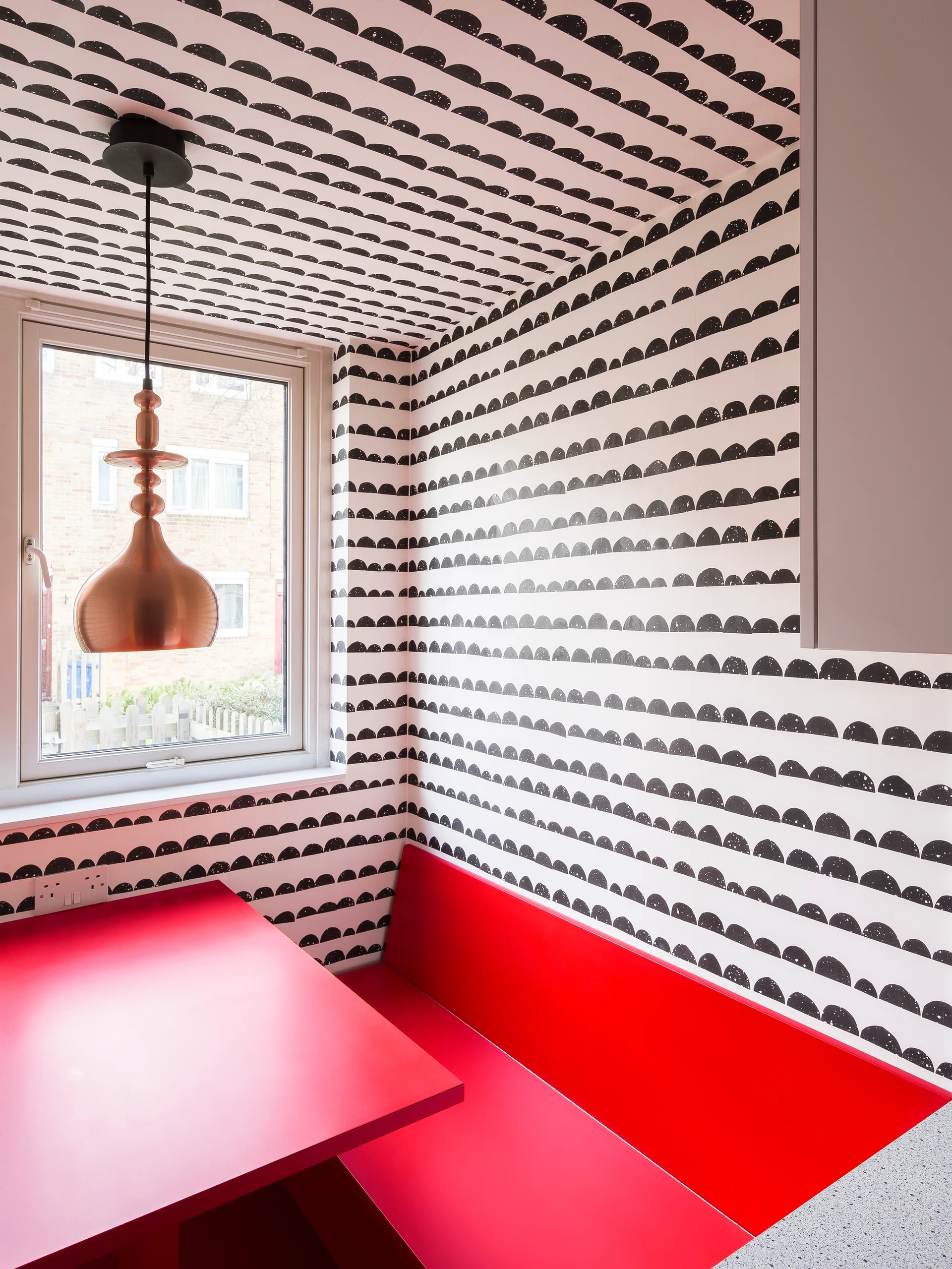 A breakfast nook with a bright red built-in bench and table set against black and white patterned wallpaper that extends to the ceiling.