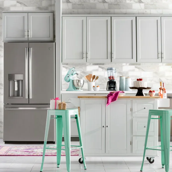 A bright kitchen features grey shaker cabinets, a white wheeled kitchen island with mint-green stools, and a stainless steel refrigerator.