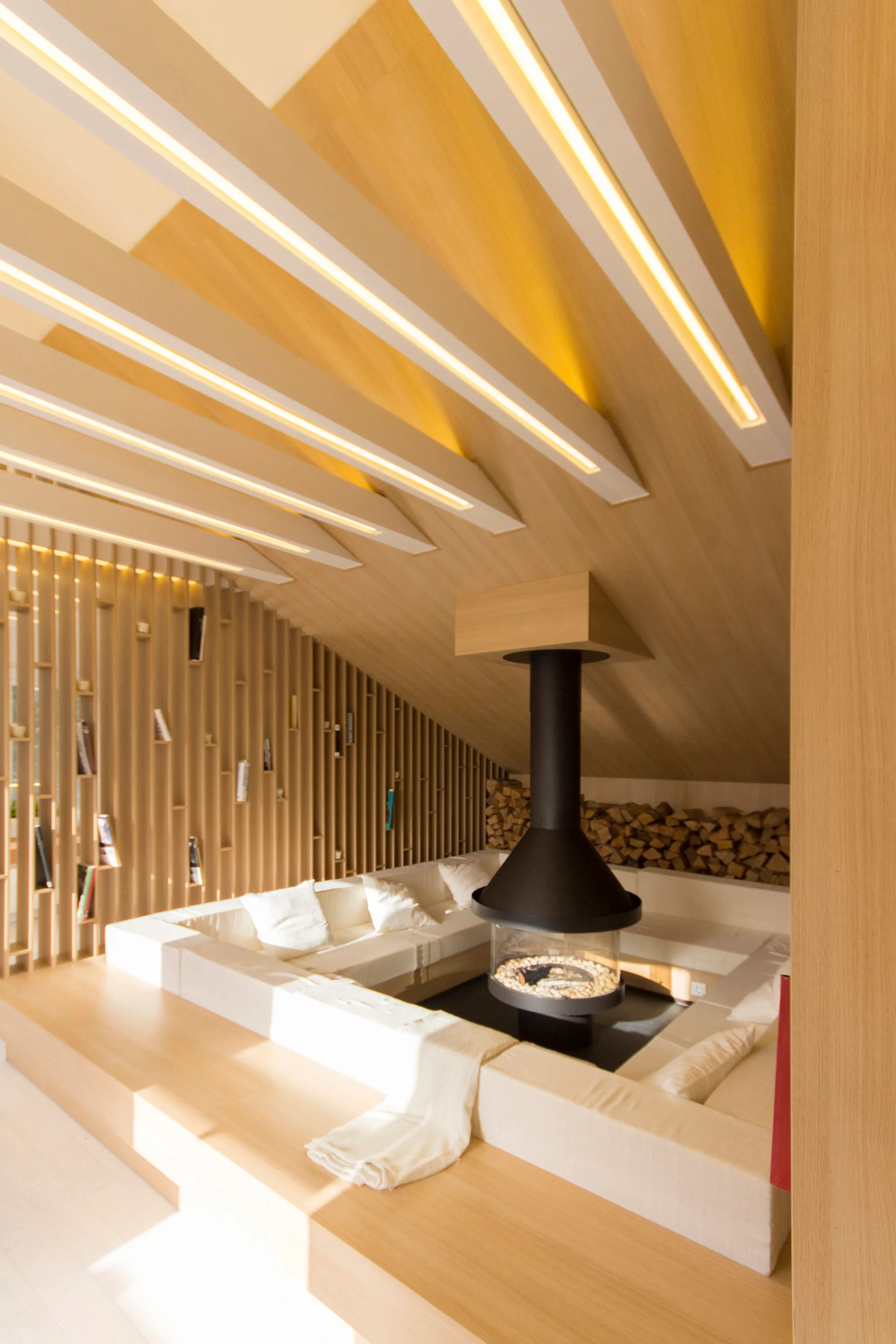 A modern sunken lounge features a central black suspended fireplace, built-in white seating, and a sloped wood ceiling with glowing integrated light beams.