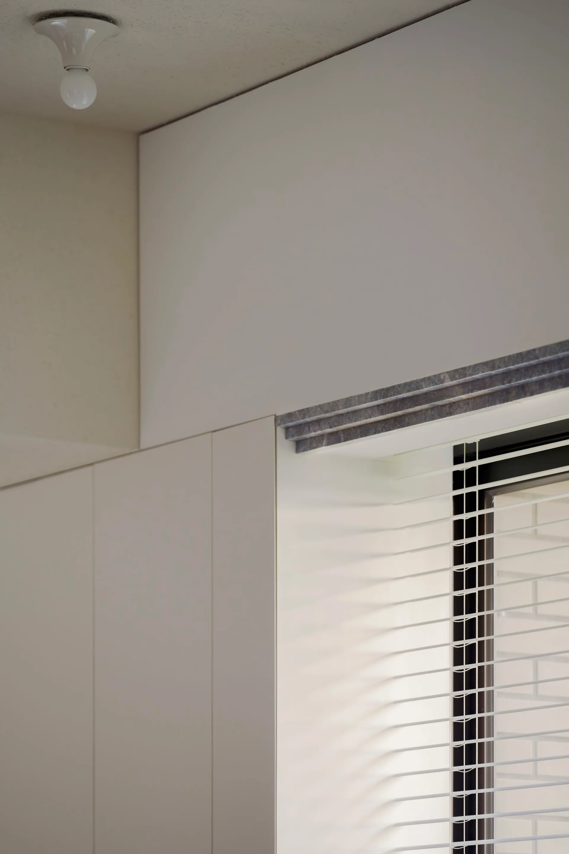 Minimalist off-white built-in cabinets stand next to a window with horizontal blinds and a textured grey stone header under a flush-mount ceiling light.