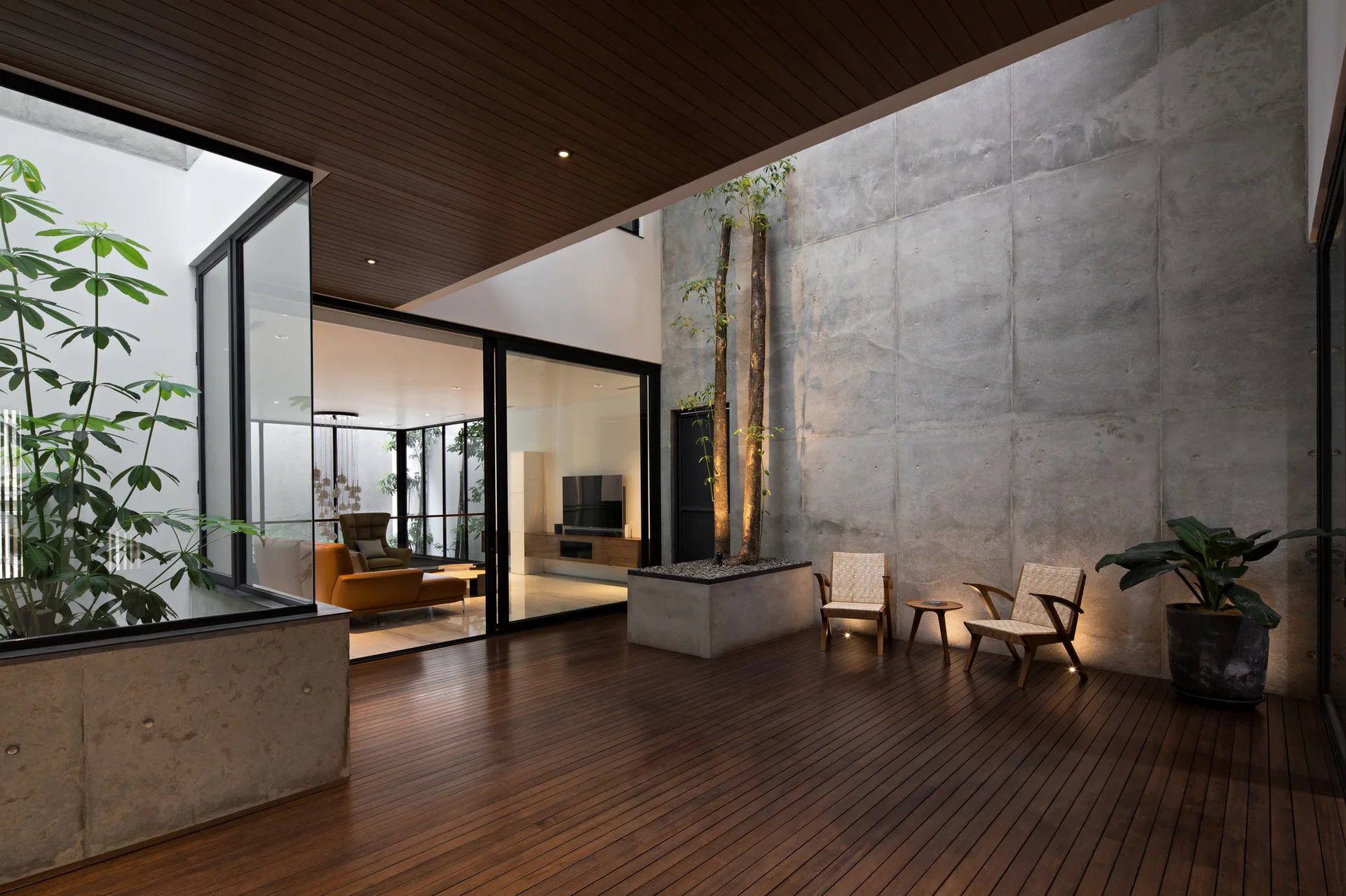 A modern patio with dark wood decking, a large concrete feature wall, two woven armchairs, and sliding glass doors showing an indoor living room.