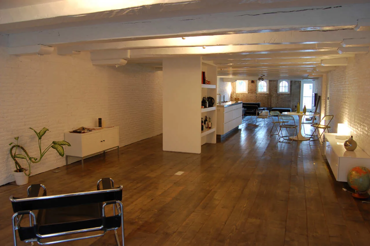 An open-plan loft apartment features painted white brick walls, exposed ceiling beams, a long hardwood floor, and minimalist furniture.
