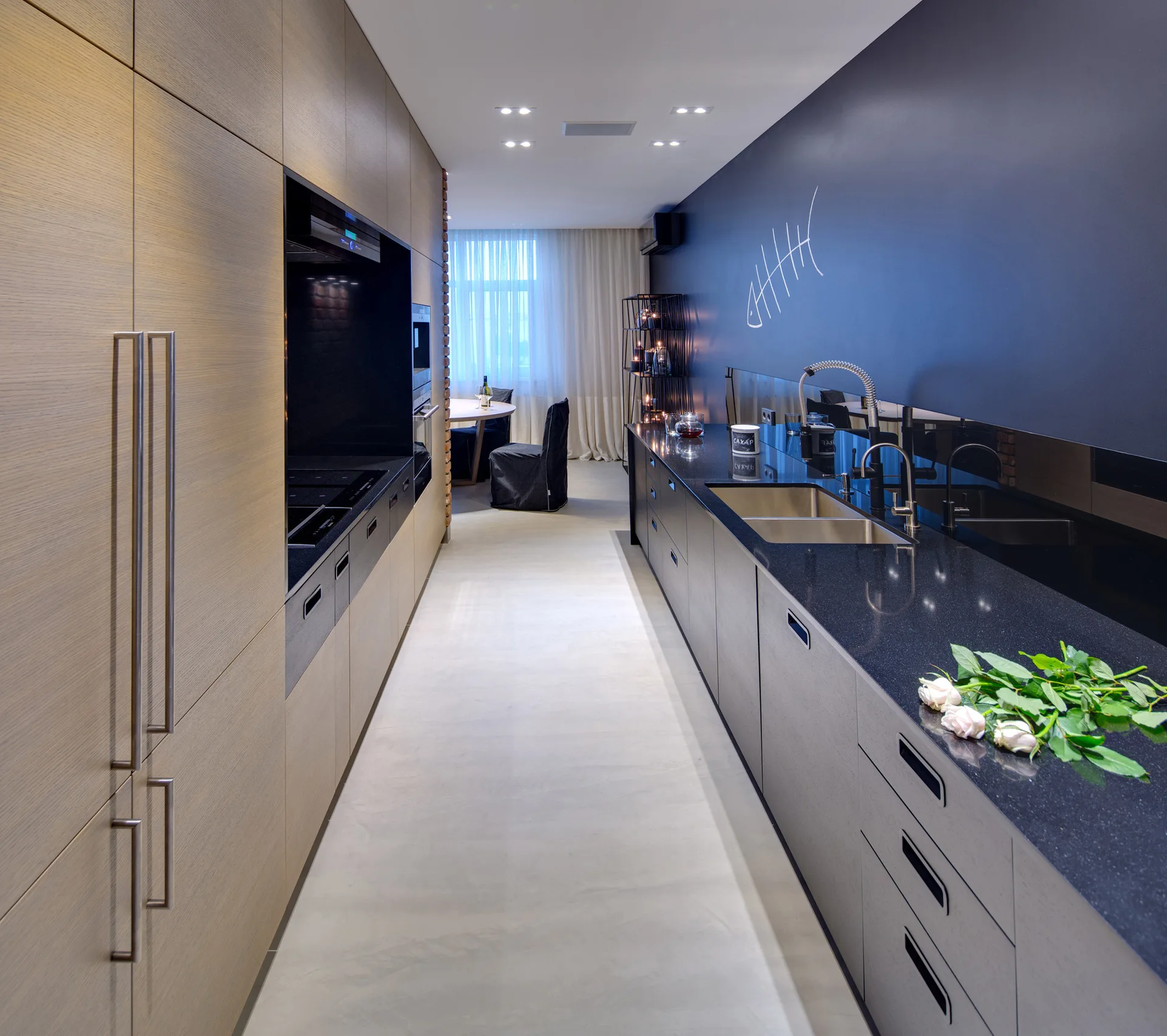 A modern galley kitchen featuring tall wood-toned cabinets, black countertops, grey lower cabinets, and a deep navy wall.