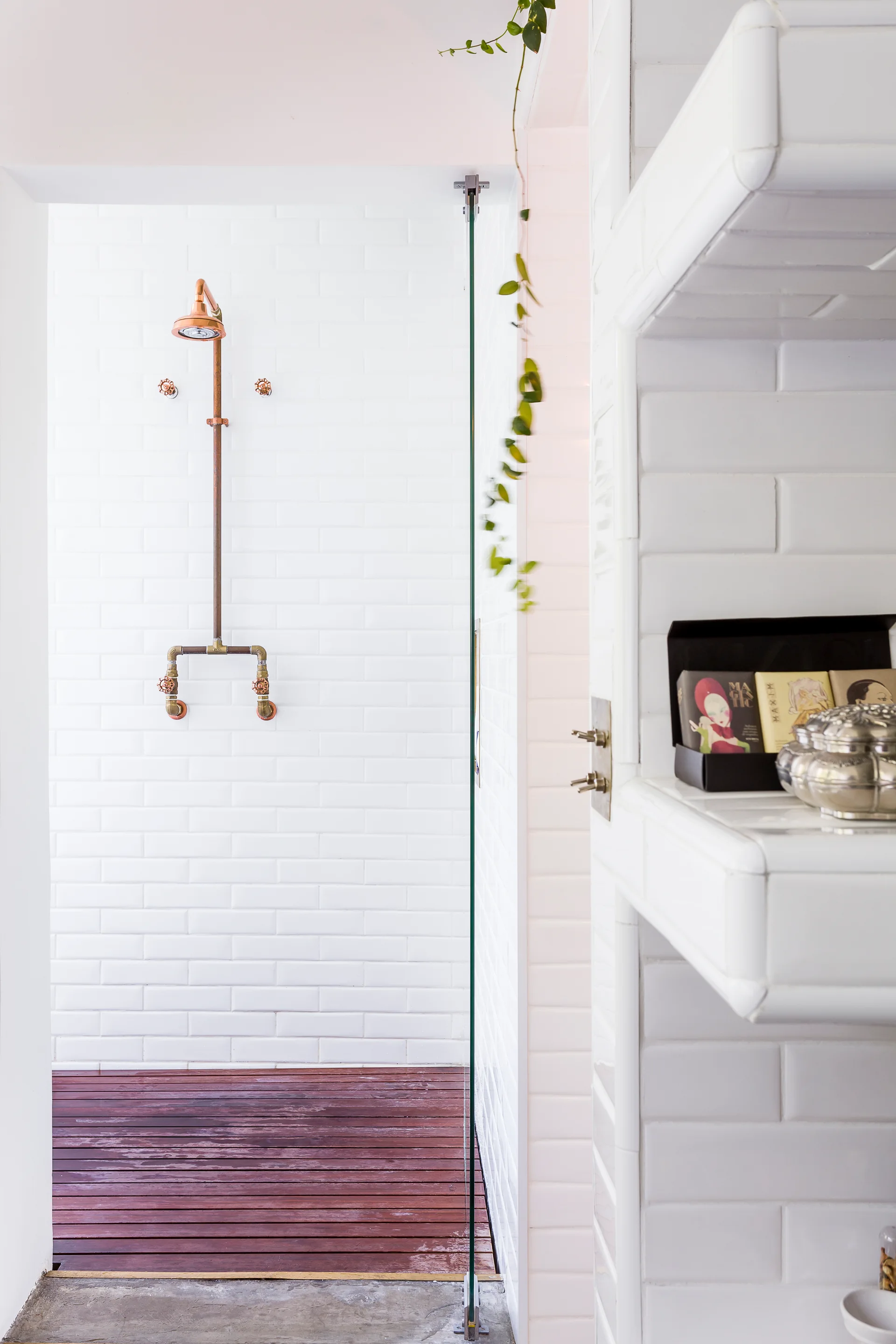 A modern walk-in shower features white subway tile walls, an exposed copper pipe plumbing fixture, and dark wood plank flooring.