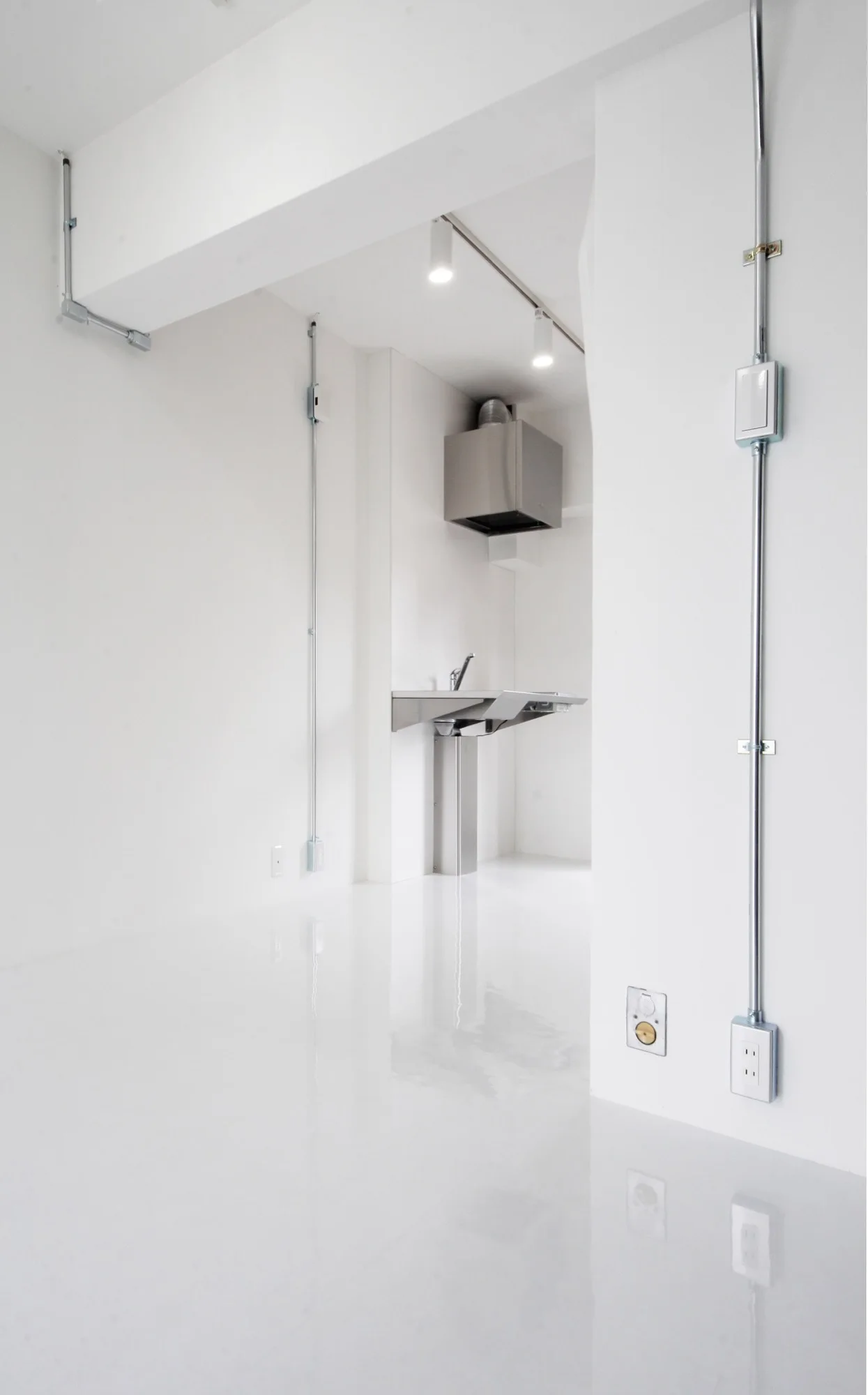 A minimalist white room features a glossy reflective floor, exposed metal electrical conduits on the walls, and a stainless steel wall-mounted sink.
