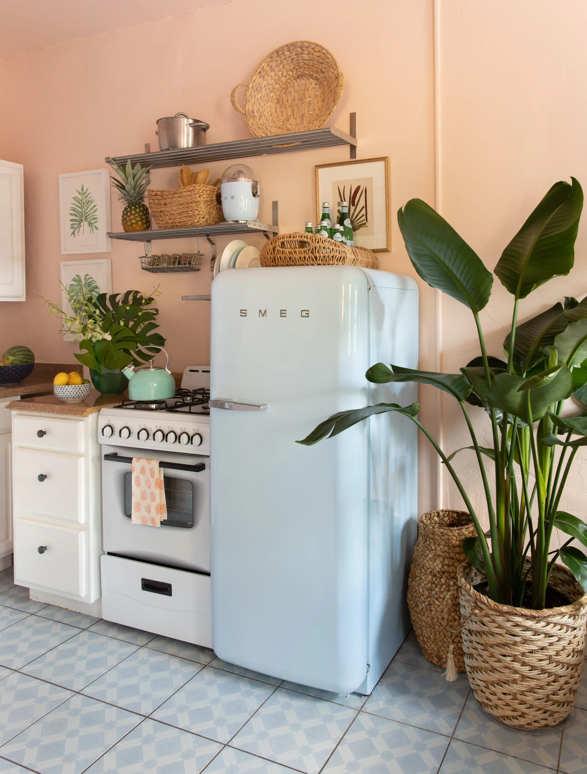 A retro kitchen with pastel pink walls, a light blue refrigerator, white cabinets, and blue and white patterned tile flooring.