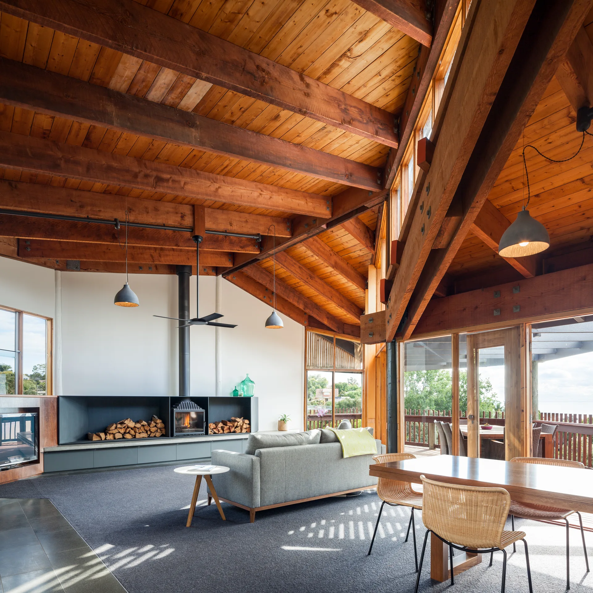 A living and dining room featuring an exposed timber-beam vaulted ceiling, a gray sofa, a black built-in fireplace, and wicker chairs.