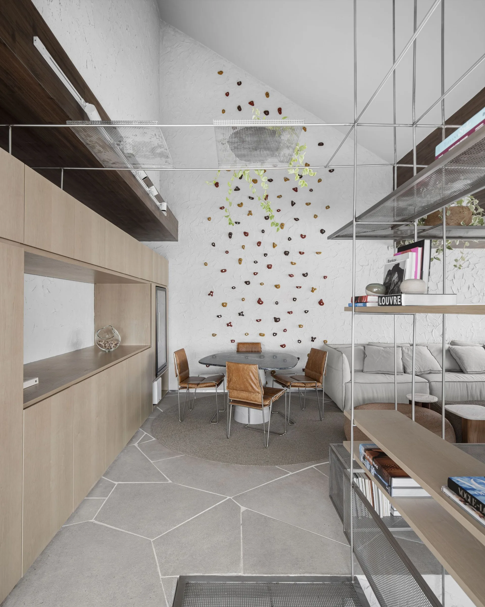 A modern dining area with a round table, leather chairs, and an indoor climbing wall built on a white textured plaster accent wall.