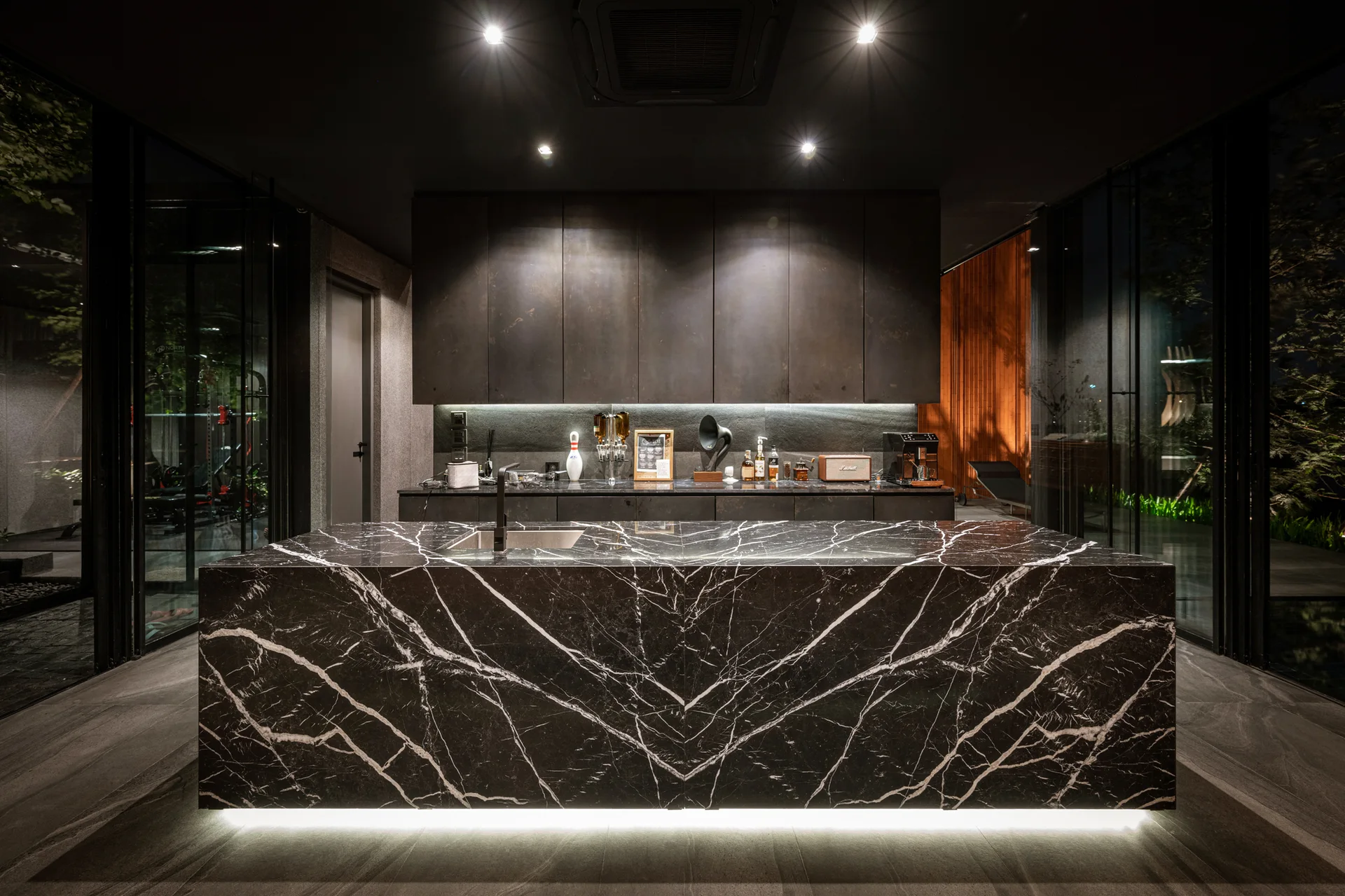 A dark modern kitchen featuring a black veined marble island illuminated by under-counter lighting, set against dark matte cabinetry.
