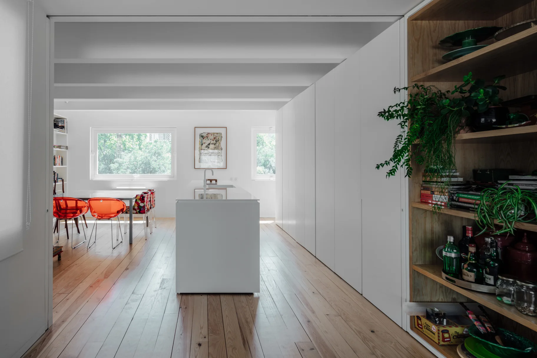 A bright minimalist kitchen features a central white island, floor-to-ceiling white cabinets, and a wood shelving unit on light-colored wood flooring.