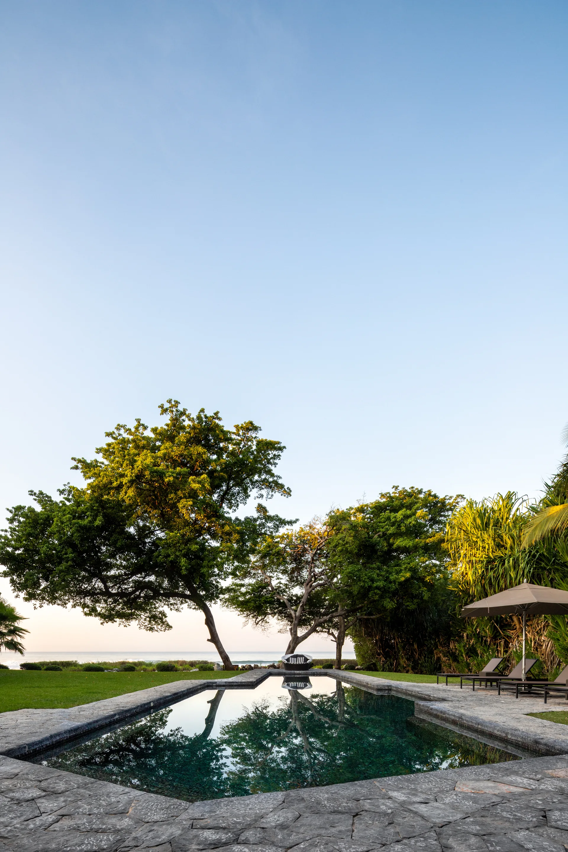 A rectangular swimming pool with dark water reflecting trees is surrounded by irregular grey stone decking and a grass lawn under a clear sky.