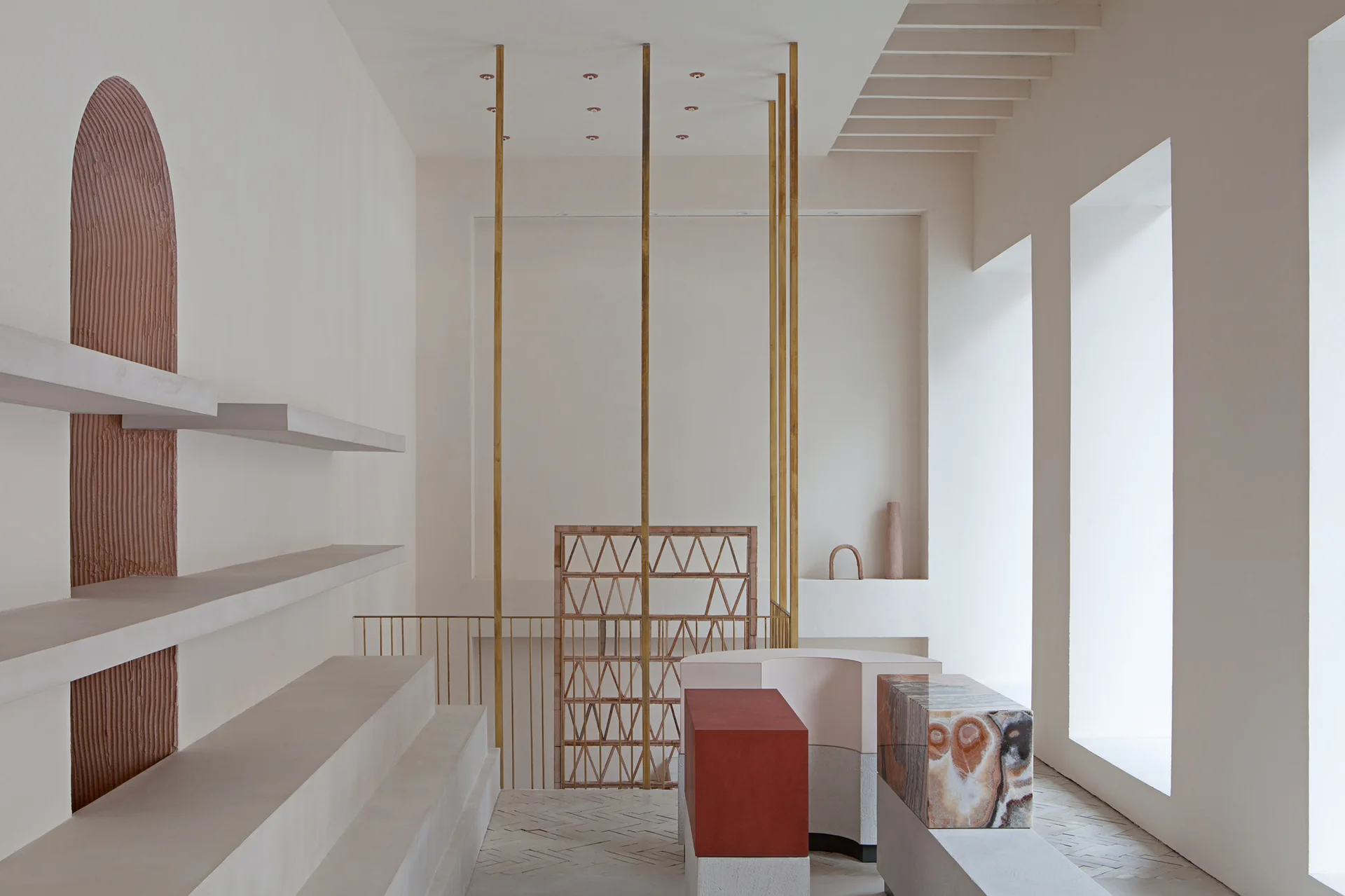 A minimalist room with built-in plaster shelving, a textured terracotta wall arch, and multi-material sculptural blocks on a herringbone floor.