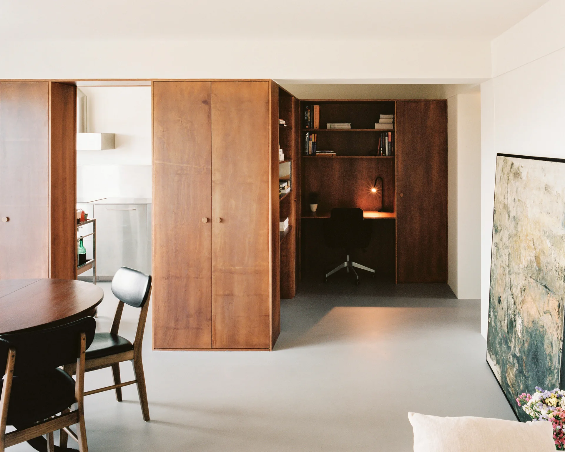 A modern interior featuring a large wood veneer partition that separates a dining area from a built-in office desk nook with grey flooring.
