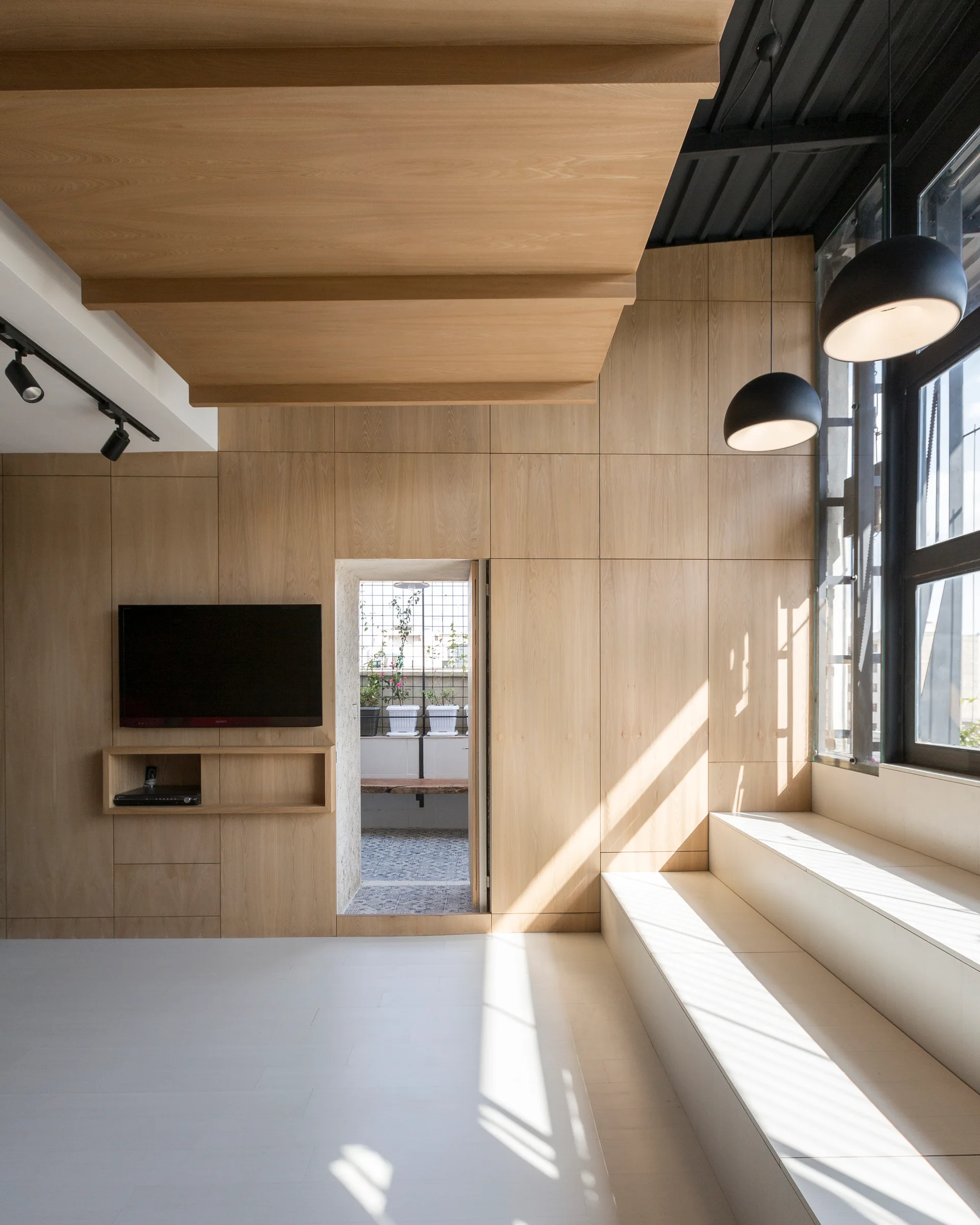 A minimalist living room features floor-to-ceiling wood paneling, a flat-screen TV, a stepped seating platform, and black pendant lights.