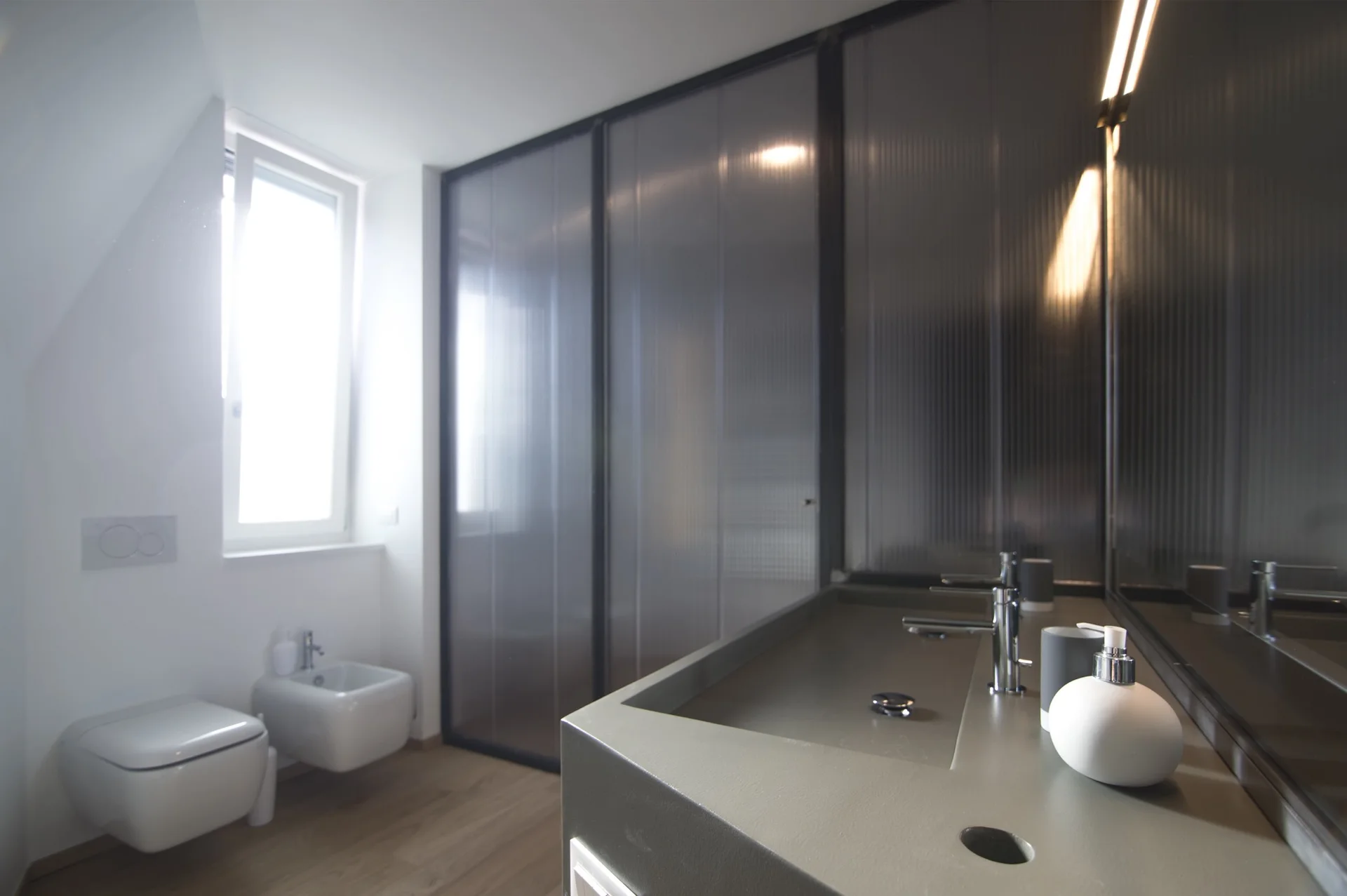 A minimalist bathroom with a gray integrated vanity, a wall-mounted toilet and bidet, and a fluted glass partition wall.