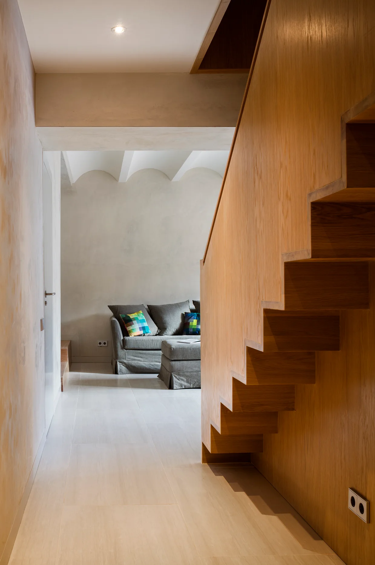 A hallway featuring a solid wood staircase with an exposed geometric profile leads to a grey sofa in a neutral living room.