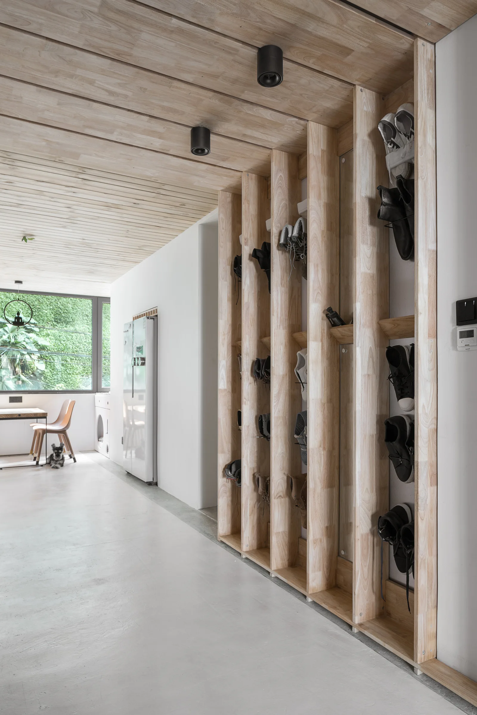 A minimalist hallway features custom vertical wooden shoe shelving, a matching wood plank ceiling, black track lighting, and a grey concrete floor.