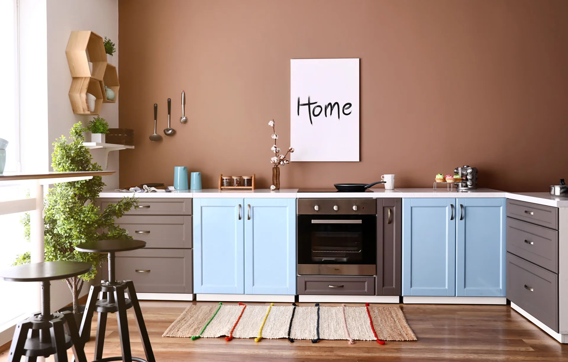 A modern kitchen featuring blue and grey base cabinets beneath a warm brown painted wall with a built-in oven and wooden floors.