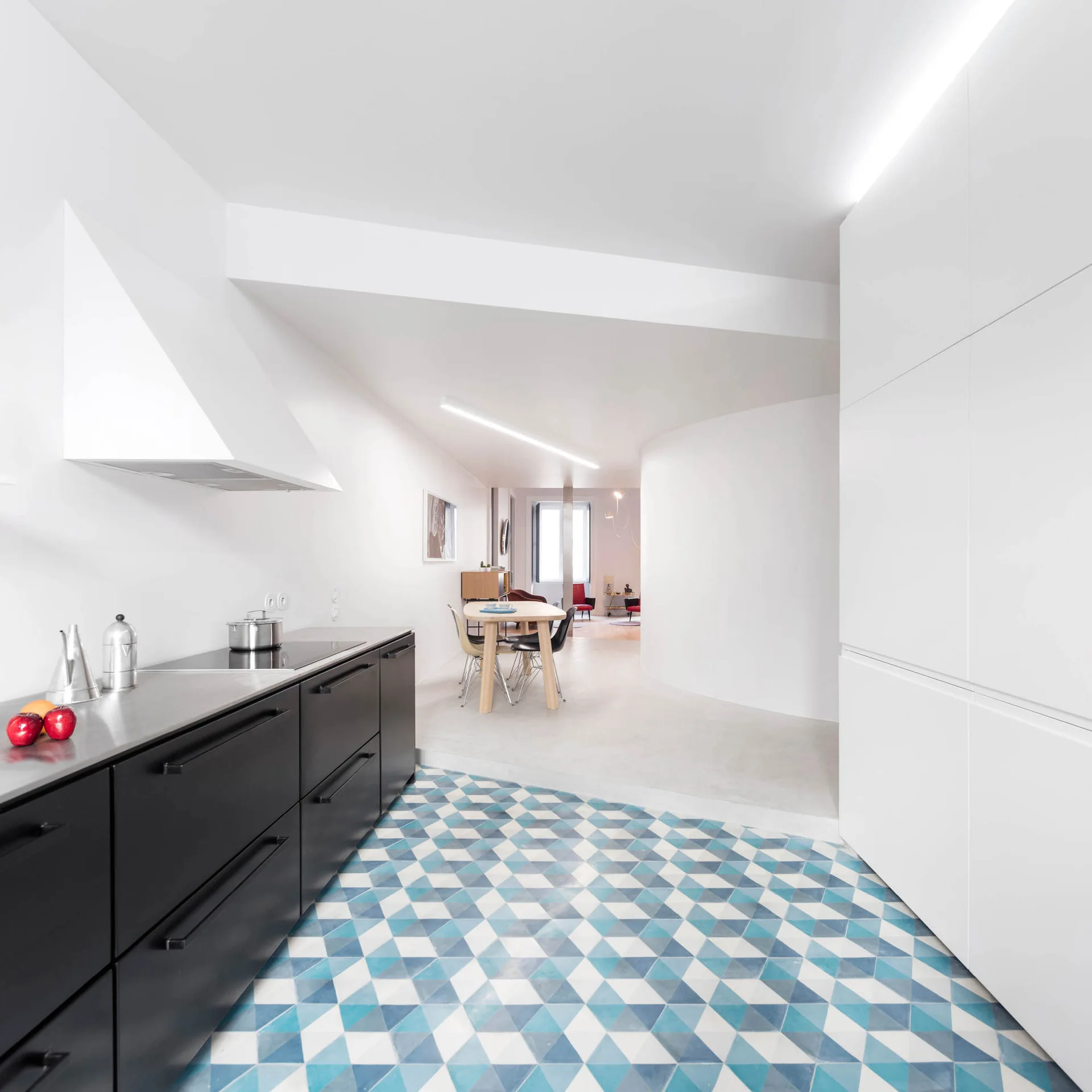 A minimalist kitchen features matte black base cabinets, white tall cupboards, and a striking blue and white geometric tile floor leading to a dining area.