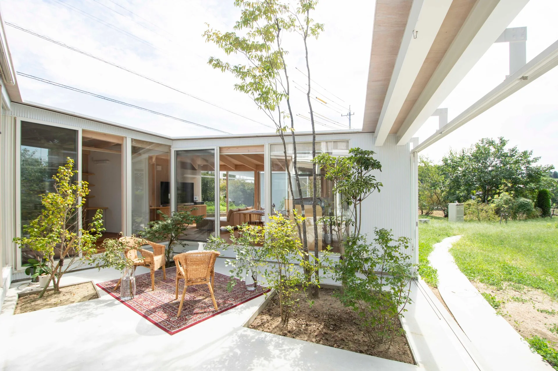 Two rattan armchairs sit on a red patterned rug on a concrete patio next to a modern glass-walled house with integrated garden planters.