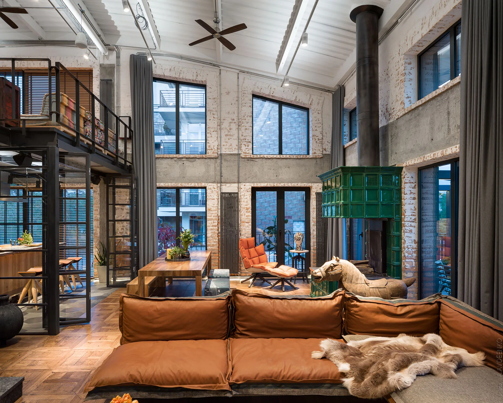 An industrial loft living room featuring a brown leather modular sofa, a tall green ceramic fireplace, and exposed brick walls.
