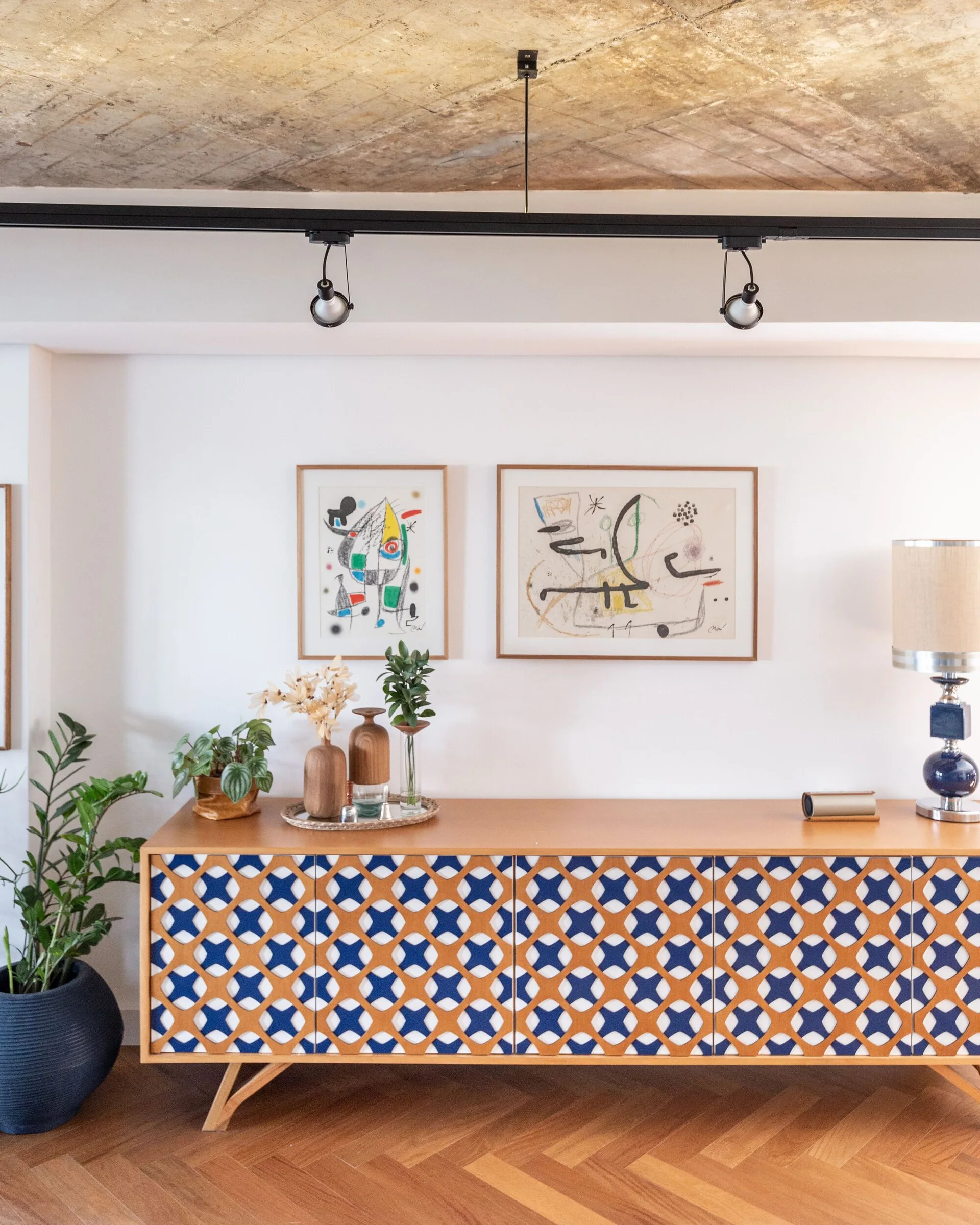 A wooden sideboard with blue-and-white patterned doors sits against a white wall under an exposed concrete ceiling with track lighting.