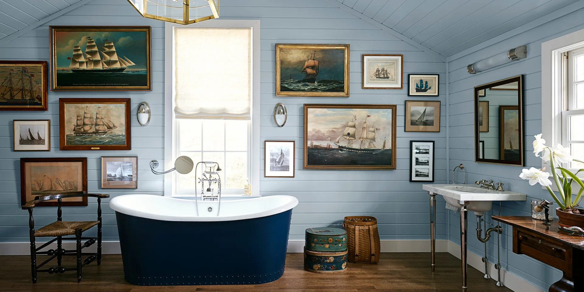 A light blue bathroom with a navy soaking tub, a marble washstand, and a gallery wall of framed maritime paintings.