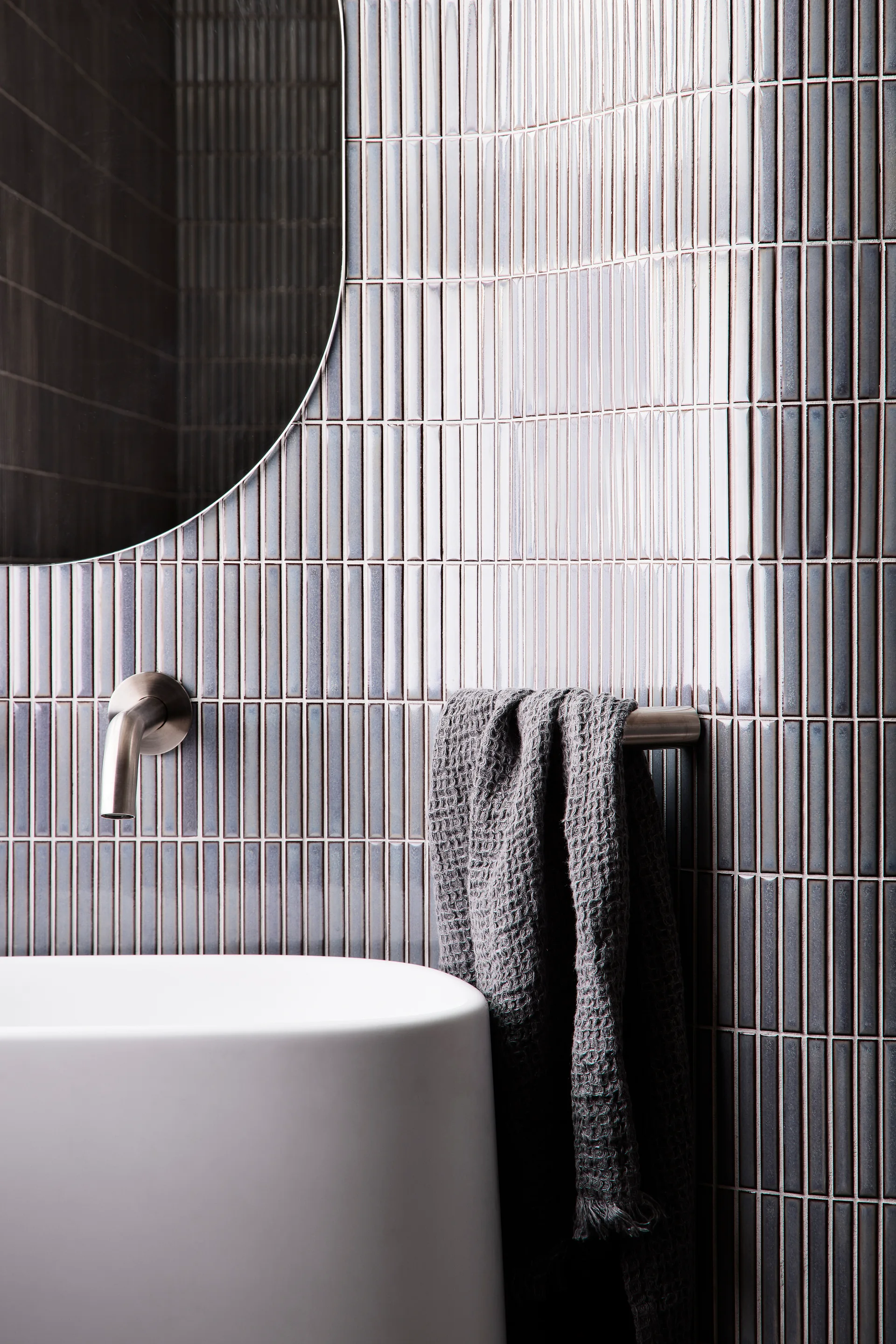 A close-up of a bathroom wall featuring grey vertically stacked kit-kat tiles, a wall-mounted silver faucet, a white washbasin, and a grey towel.