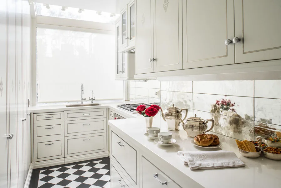 A bright white kitchen with a mirrored tile backsplash, white cabinets with silver trim, and a black and white checkered floor.