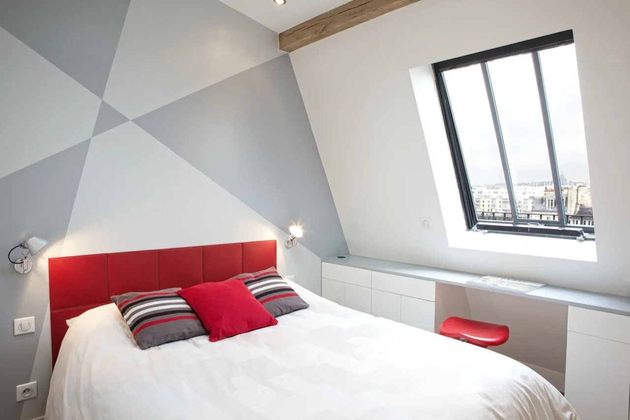 A modern attic bedroom features a bed with a red headboard, a grey geometric accent wall, and a built-in white desk under a sloped skylight window.