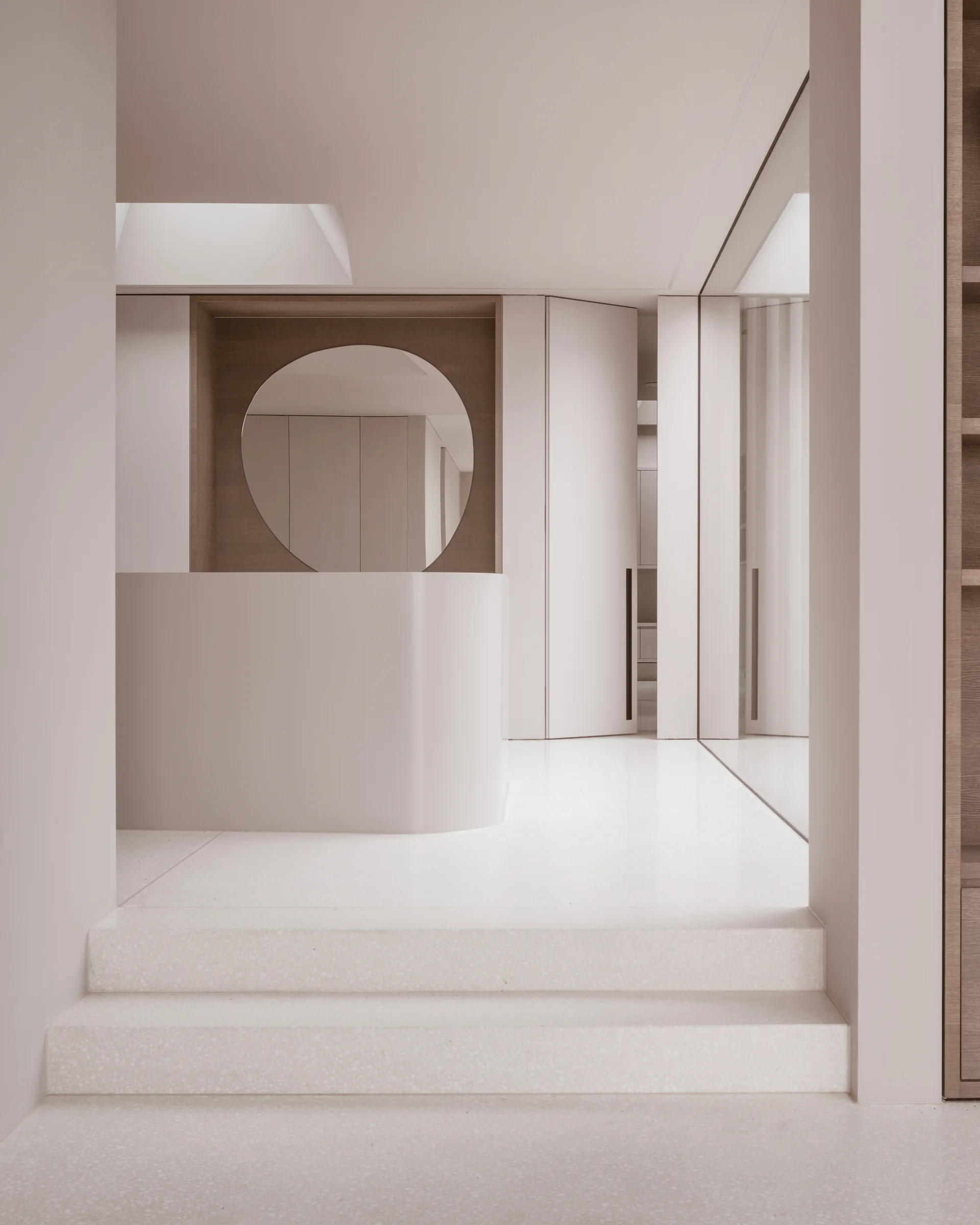 A minimalist cream entryway with speckled stone stairs leading up to a curved console table and a round mirror set in a wood niche.