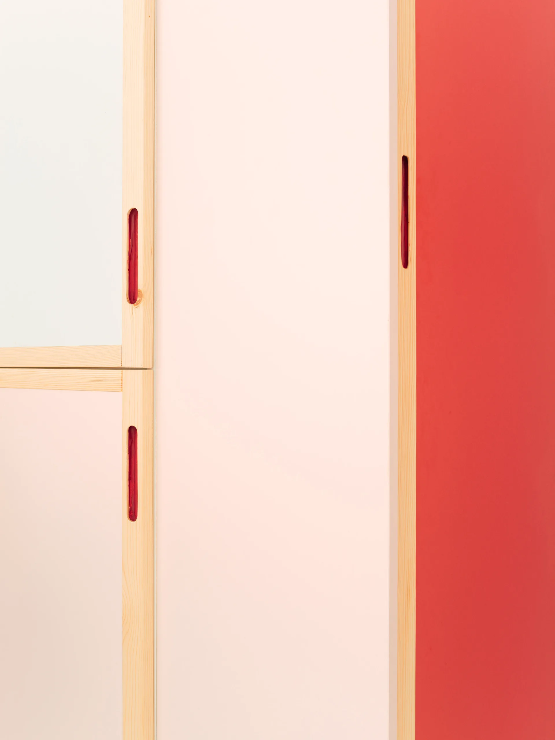 A close-up of custom cabinetry featuring matte light pink and bold red panels framed in light natural wood with carved vertical finger pulls.
