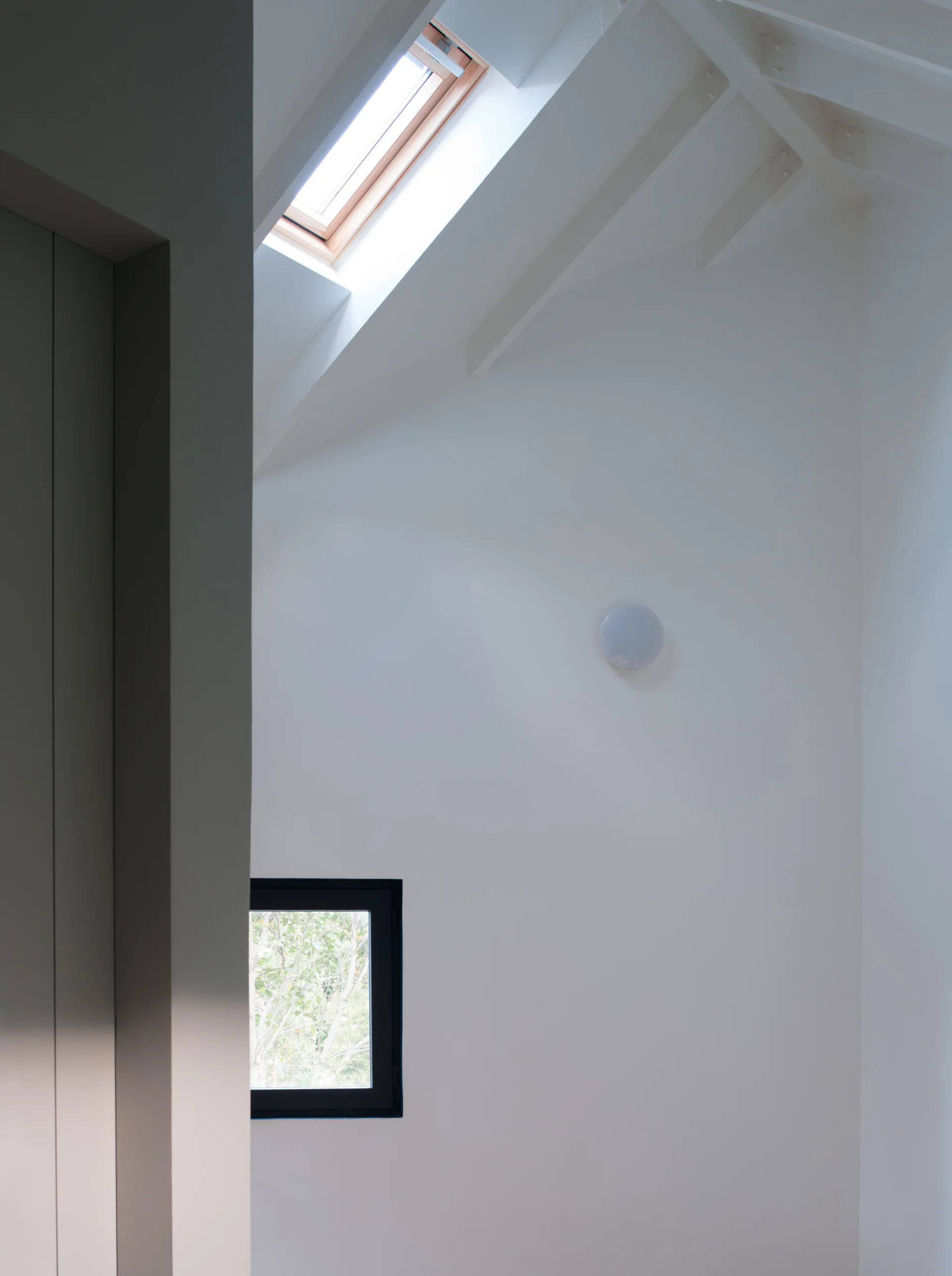 A minimalist white attic interior featuring a slanted ceiling with a wood-framed skylight and a small black-framed square window.