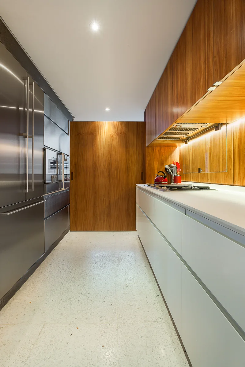 A modern galley kitchen featuring sleek white base cabinets, warm wood upper cabinetry, integrated stainless steel appliances, and a speckled floor.