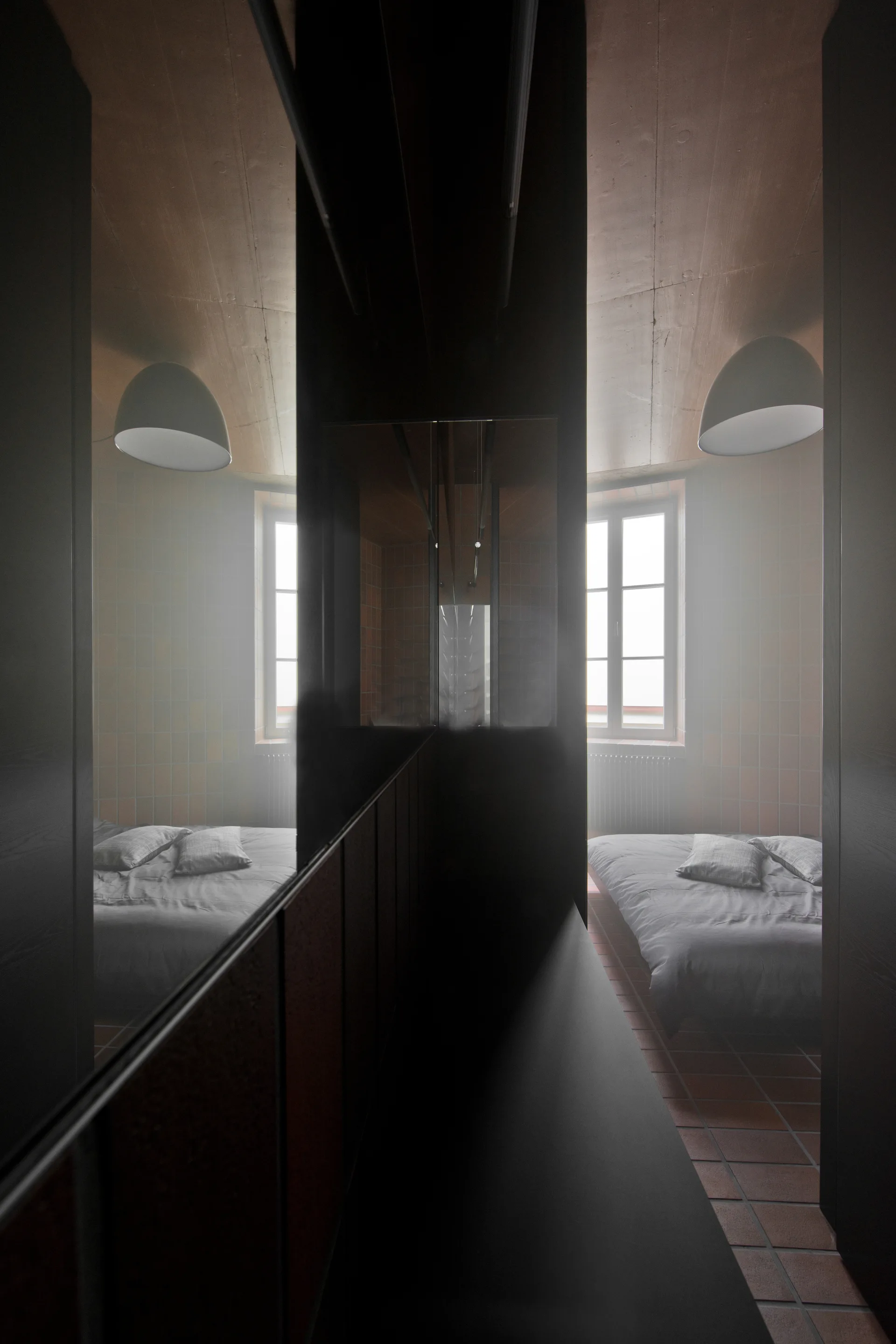 A bedroom with terracotta floor and wall tiles features a low bed and a dark, reflective room divider reflecting the room and window.