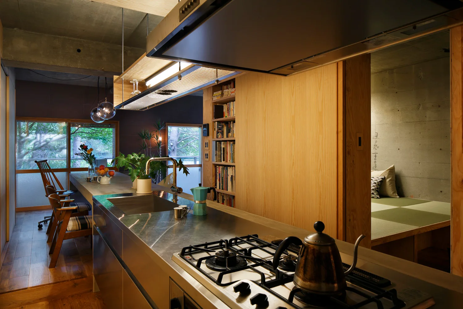 A modern kitchen featuring a stainless steel island, an integrated wooden dining table, and a built-in bookshelf next to a raised tatami niche.